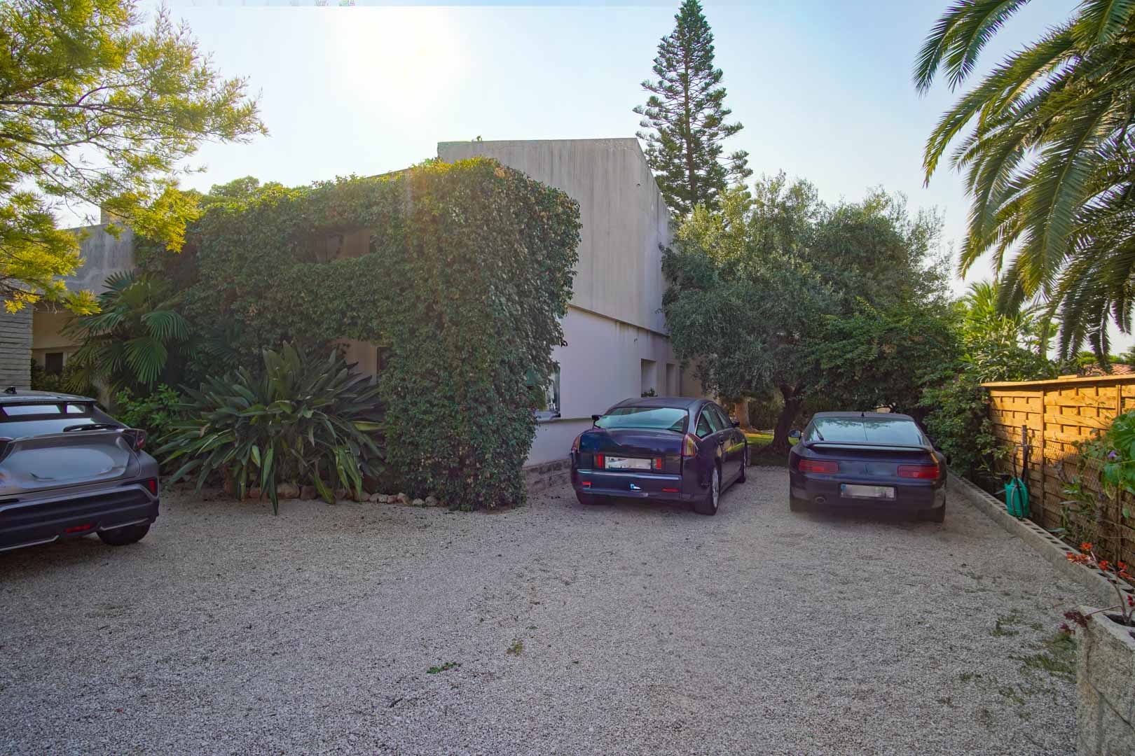 Buitenparkeerplaats met grindoppervlak, omgeven door volgroeide mediterrane beplanting en een houten omheining. Ruimte voor meerdere voertuigen op eigen terrein.