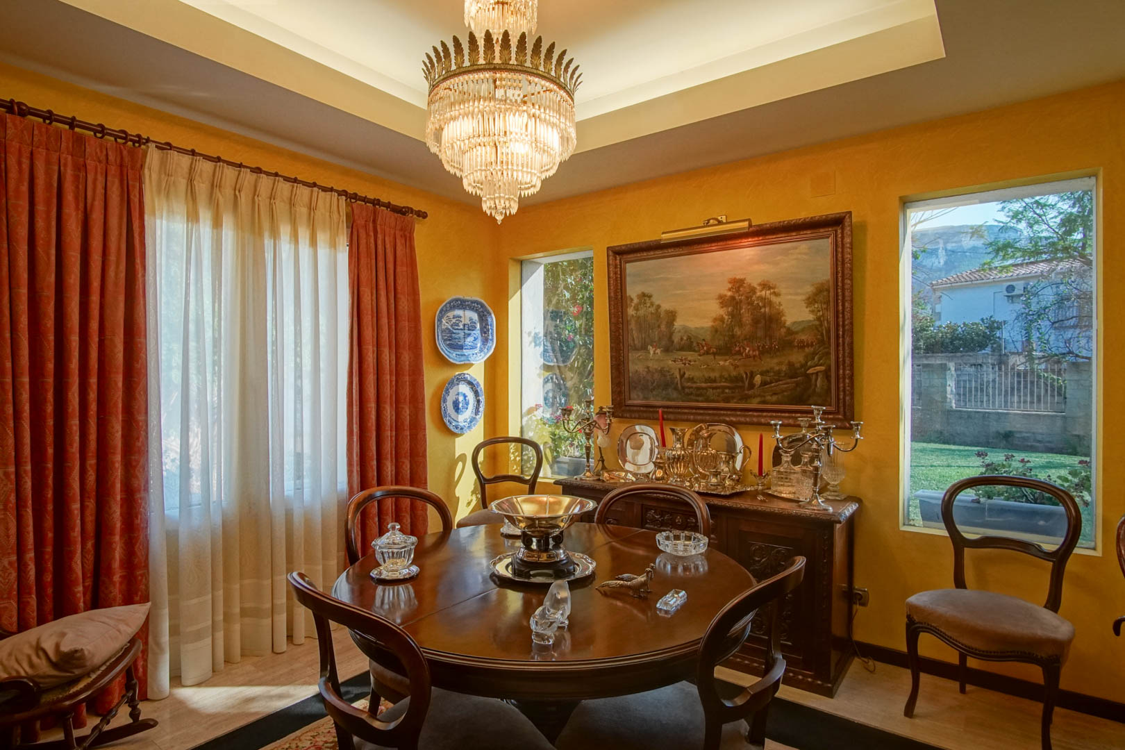 Elegante eetkamer met ronde houten tafel, kristallen kroonluchter en klassiek dressoir. De okergele muren, het koofplafond met verlichting en zicht op de tuin sieren de ruimte.