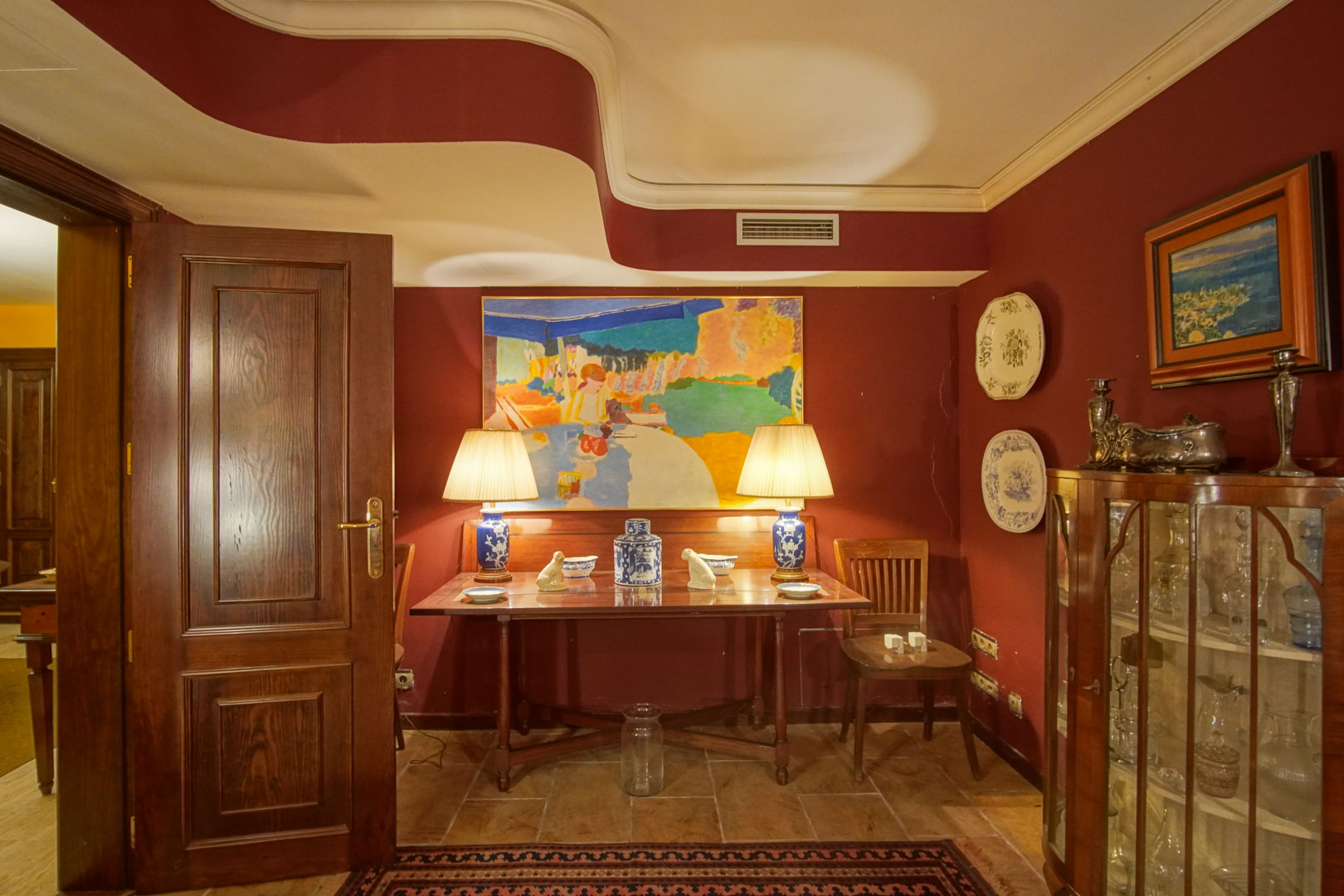 Elegante hoek met bordeauxrode muren, een uniek golvend plafond en klassiek houten meubilair. Bevat een glazen vitrinekast en keramische lampen op een natuurstenen vloer.