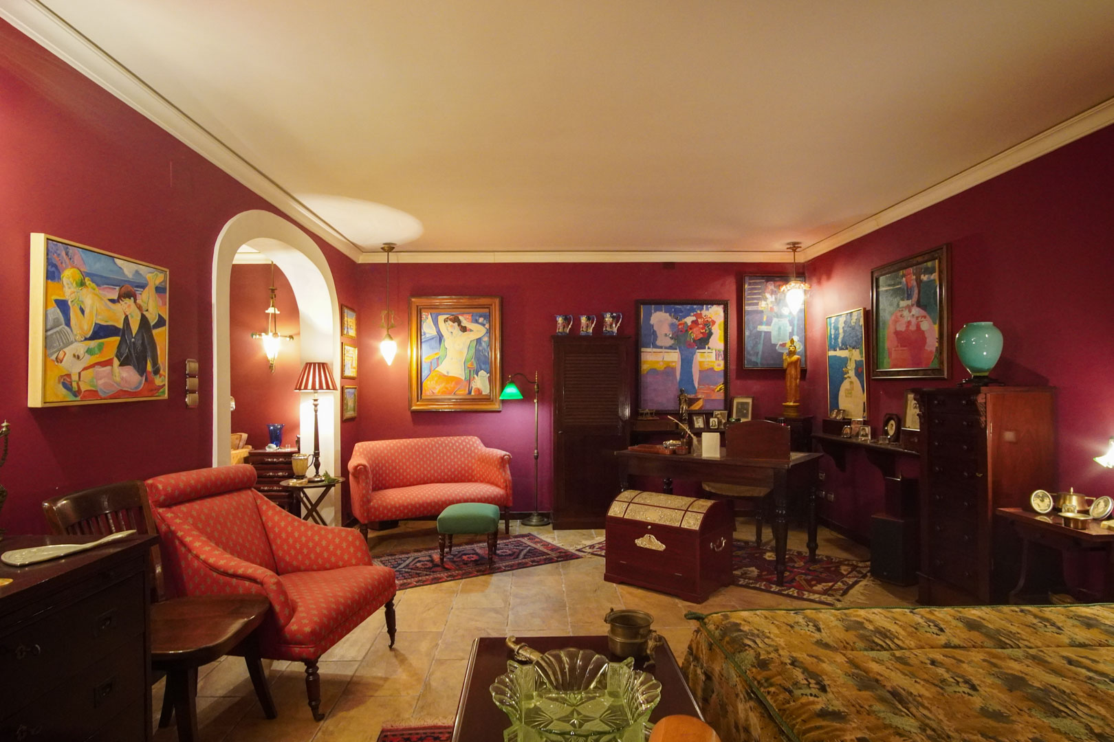 Klassieke woonkamer met bordeauxrode muren, marmeren vloeren en een werkhoek. De ruimte is ingericht met kunstcollecties, antiek meubilair en sfeervolle verlichting.