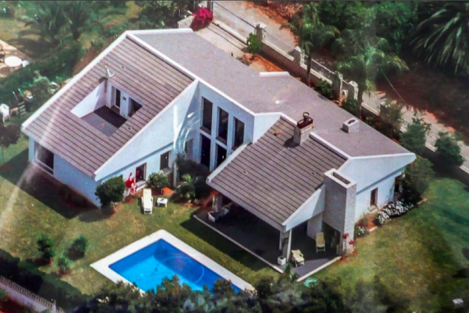Luchtfoto van de witte villa met pannendak, rechthoekig zwembad en verzorgde tuin met overdekt terras en omheining voor optimale privacy.