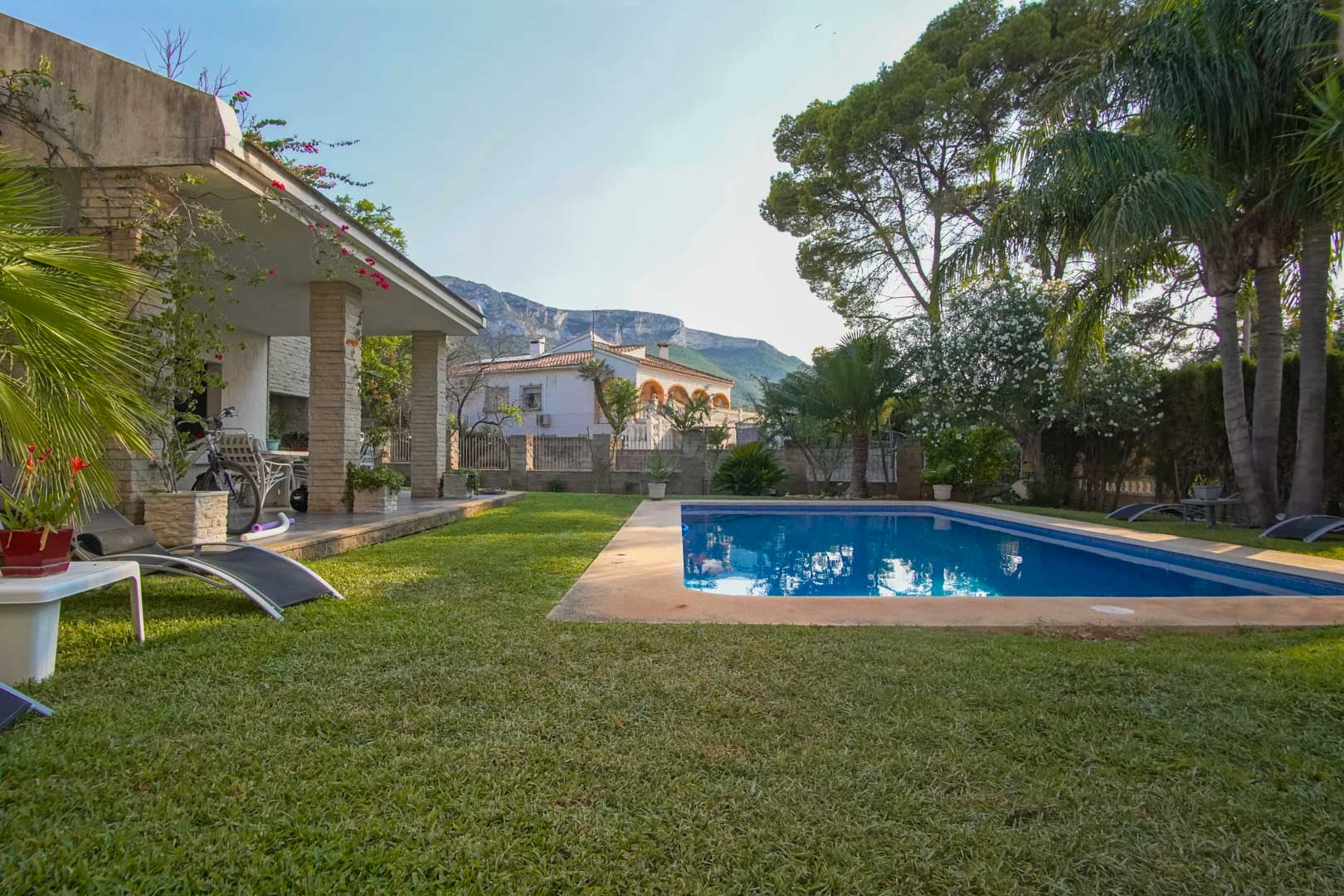 Mediterrane tuin met natuurlijk gazon, rechthoekig zwembad en overdekt terras met stenen zuilen. Op de achtergrond is het silhouet van de Montgó-berg zichtbaar.