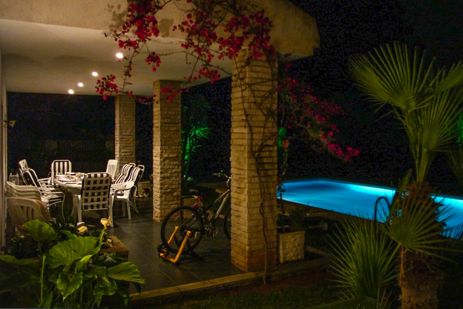 Overdekt terras met bakstenen kolommen, eethoek buiten en bougainvillea naast een verlicht zwembad in een sfeervolle nachtelijke setting.