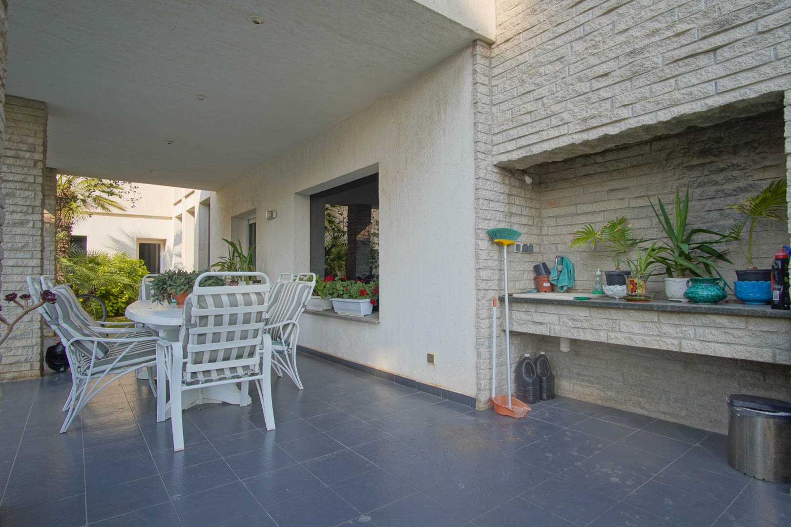 Overdekt terras met donkere tegelvloer, een eethoek buiten en een ingebouwde stenen buitenkeuken voorzien van een spoelbak en werkblad.