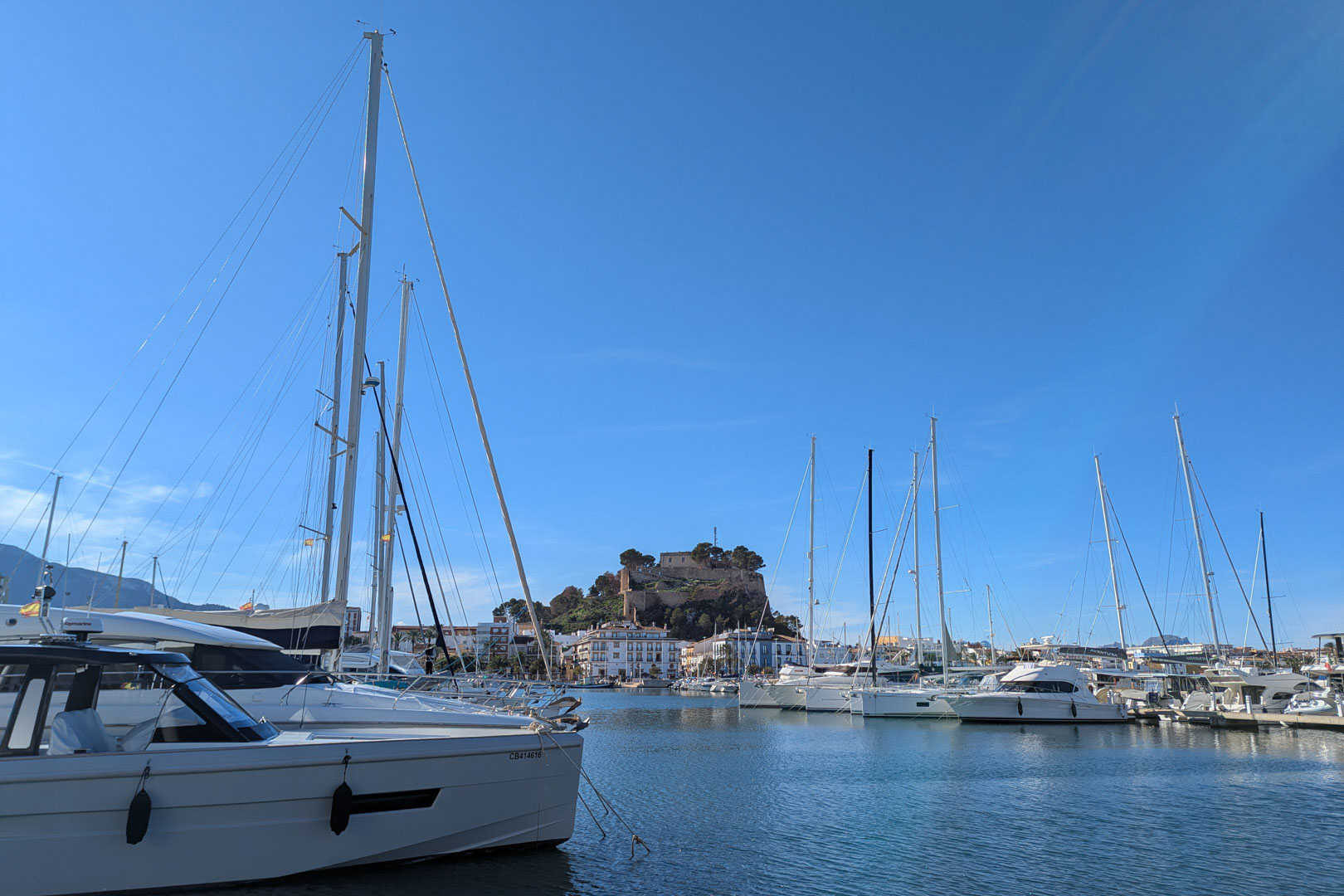 Panoramisch uitzicht op de jachthaven van Dénia met luxe jachten en het historische kasteel dat boven de stad uittorent bij een helderblauwe hemel.