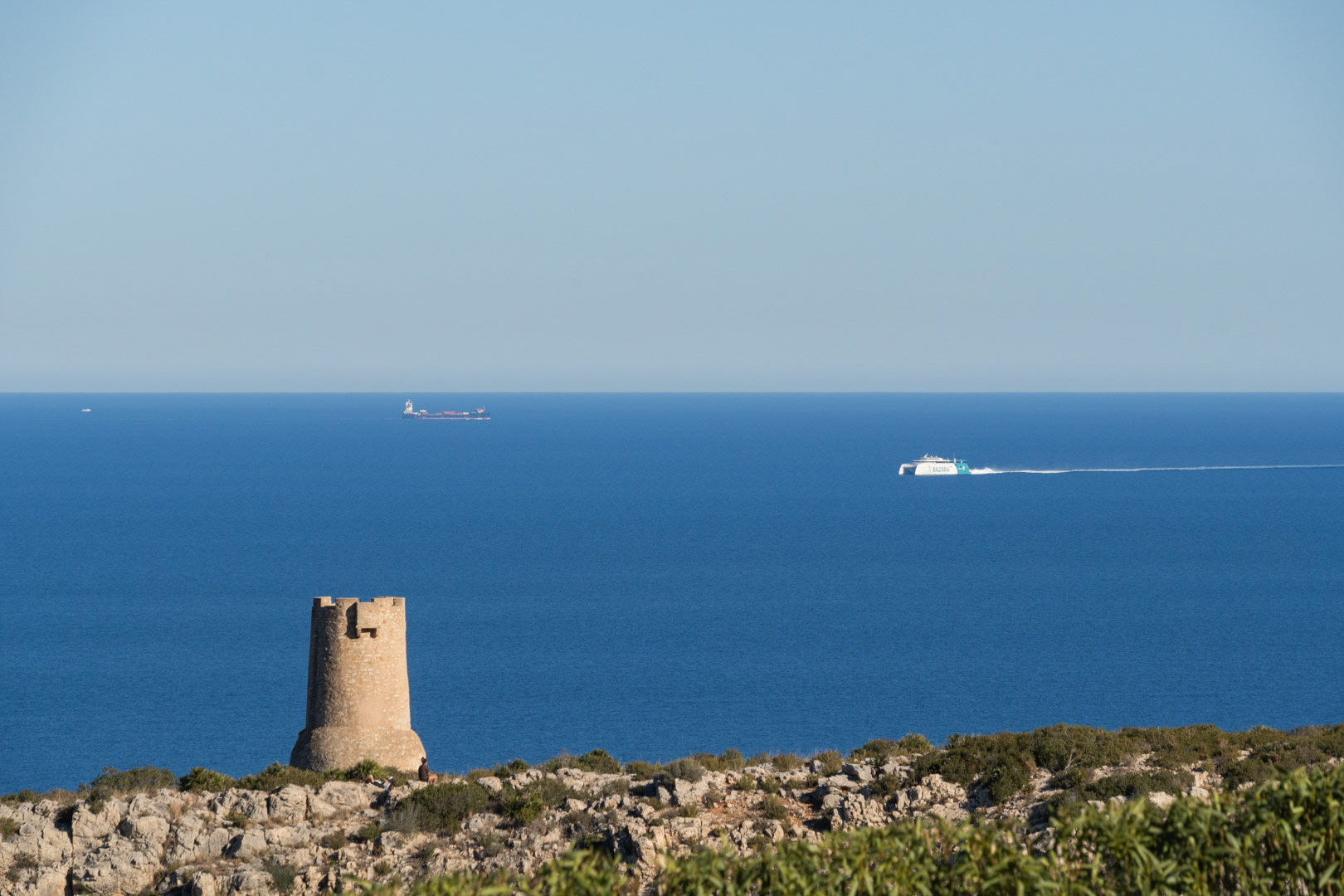 Panoramisch uitzicht op de kustlijn van Dénia met de historische Torre del Gerro-wachttoren en veerboten die over de Middellandse Zee varen.