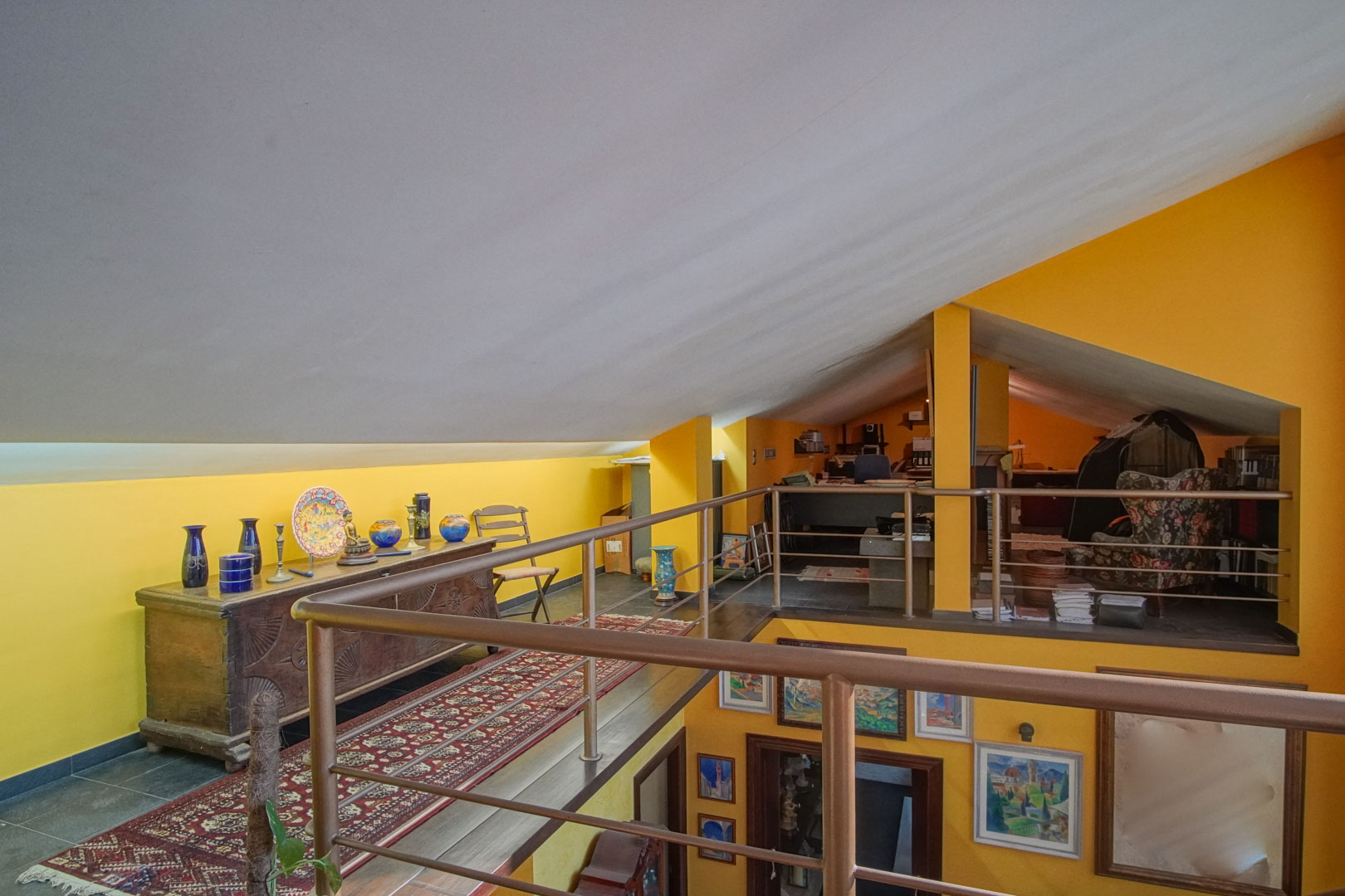 Ruime mezzanine op de bovenverdieping met schuine plafonds, gele wanden en metalen balustrades. De open ruimte is ingericht met antiek meubilair en kijkt uit op de benedenverdieping.