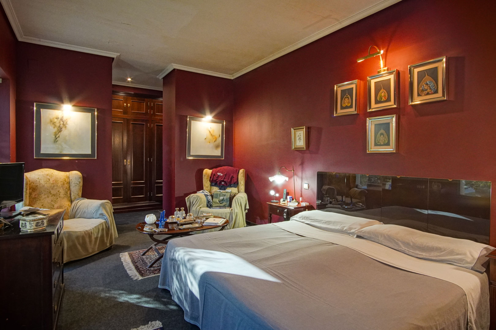 Slaapkamer met bordeauxrode muren, een zithoek met fauteuils en donkerhouten inbouwkasten. Accentverlichting accentueert de kunstwerken en de ruime indeling.