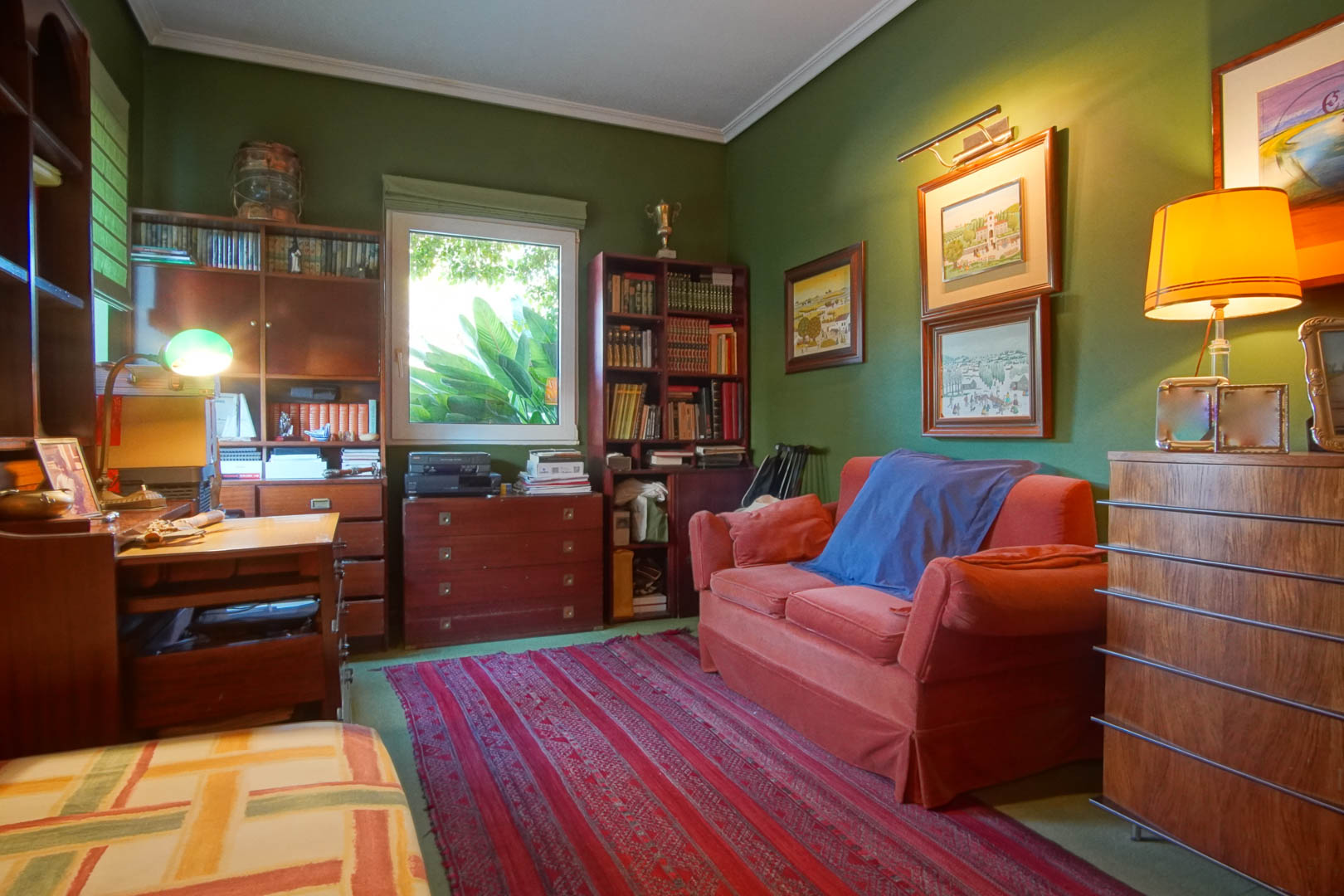 Werkkamer met groene muren en klassiek houten meubilair. Voorzien van boekenkasten, een bureau, een rode bank en natuurlijk licht met uitzicht op de tuin.