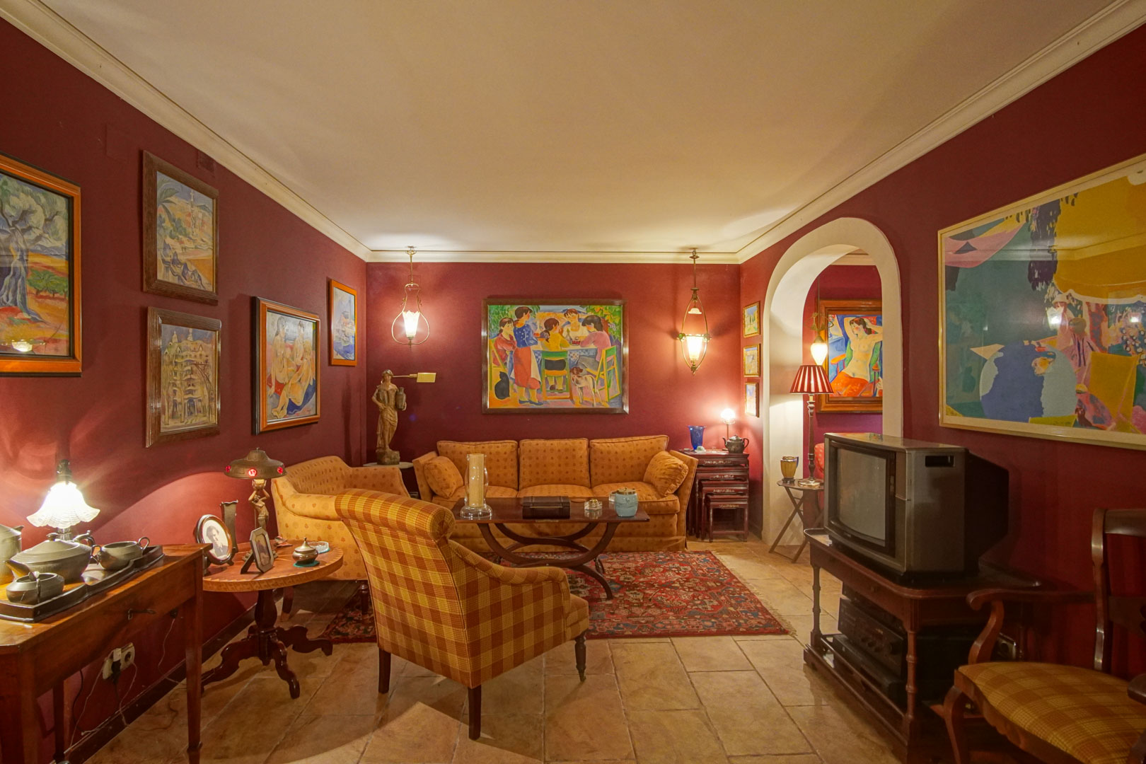 Woonkamer met bordeauxrode muren, rustieke tegelvloer en klassiek meubilair. De ruimte is voorzien van diverse kunstwerken, warme verlichting en een karakteristieke boogdoorgang.