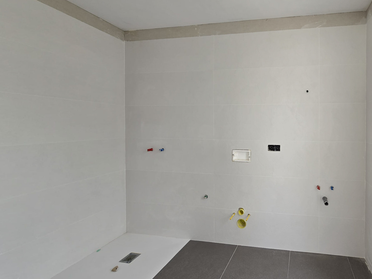 Badkamer in aanbouw met grote witte wandtegels, een drempelloze douchebak en voorbereide leidingen voor het sanitair en de elektrische voorzieningen.