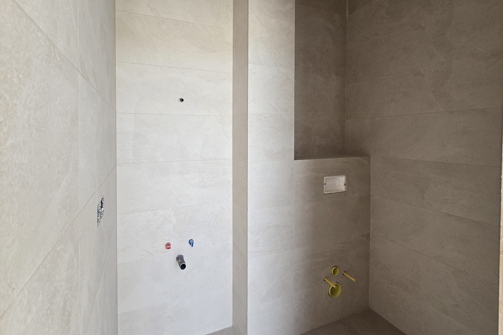 Badkamer in aanbouw met zandkleurige porseleinen wandtegels, aansluitingen voor een wastafel en inbouwreservoir voor een hangend toilet met nis.