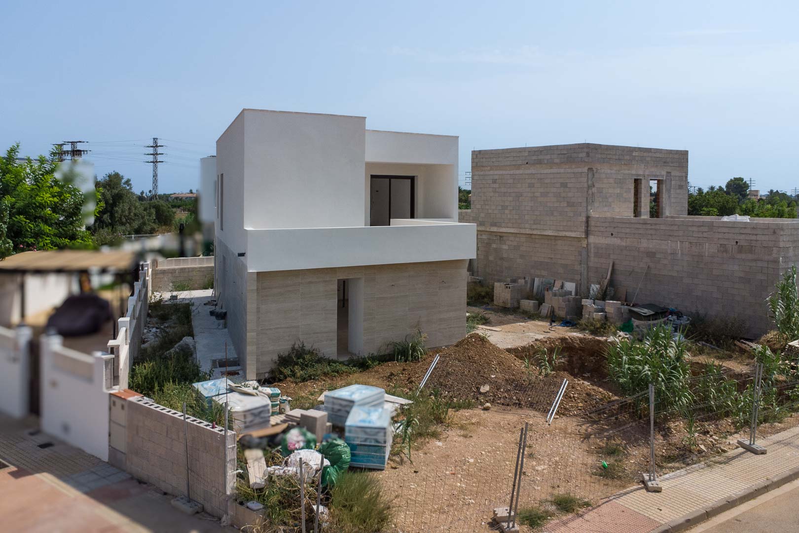 Buitenaanzicht van de villa in aanbouw met stenen gevel op de begane grond en witte bovenverdieping. De uitgraving voor het privézwembad is momenteel zichtbaar.