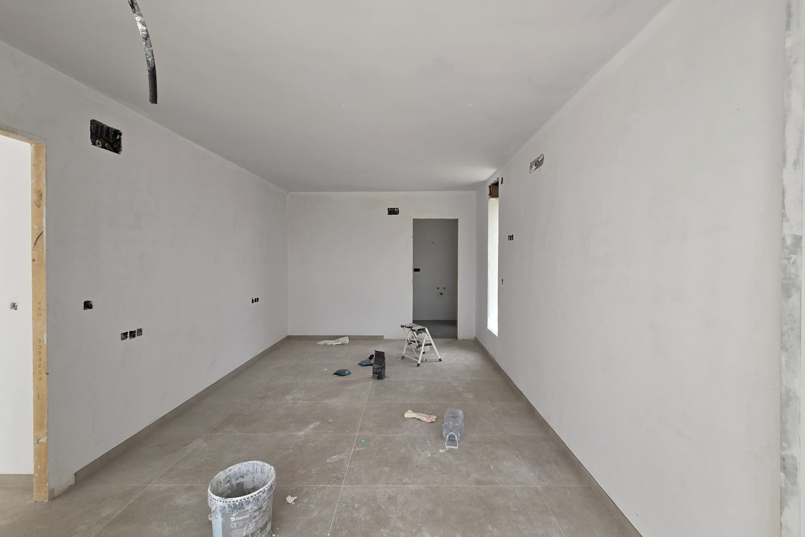Interieur in afbouwfase met grootformaat porseleinen vloertegels en gladde witte muren. Zichtbare elektrische voorinstallaties en doorgangen naar aangrenzende kamers.