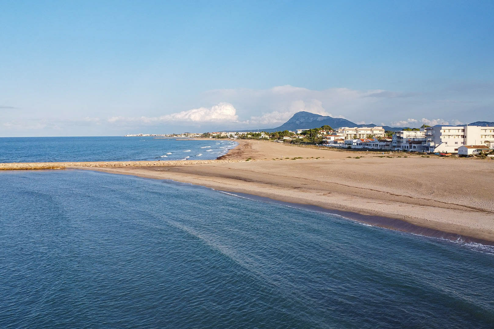 Panoramisch uitzicht op het brede zandstrand nabij de woning, met een stenen pier, de Middellandse Zee en het silhouet van de Montgó op de achtergrond.