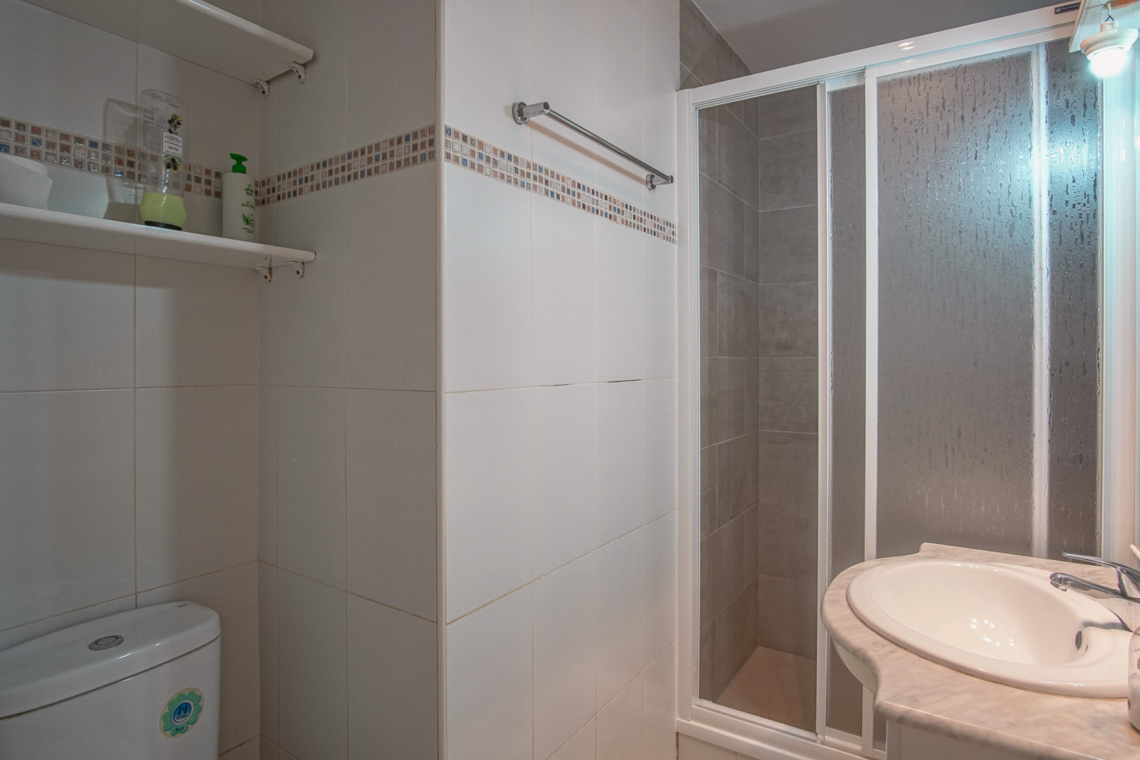 Badkamer met wit betegelde wanden en decoratieve rand, voorzien van een douchecabine met schuifdeur en een wastafel ingebouwd in een marmeren blad.