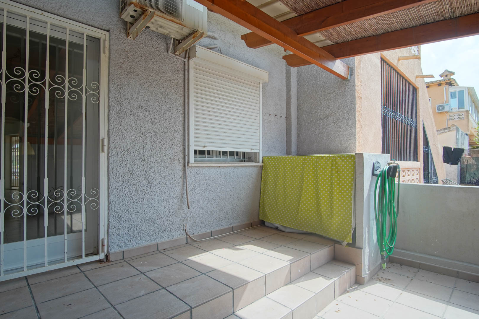 Buitenterras met tegelvloer, houten pergola met rieten dak en beveiligde toegangsdeur. Voorzien van een airconditioningunit en een praktische buitenruimte.