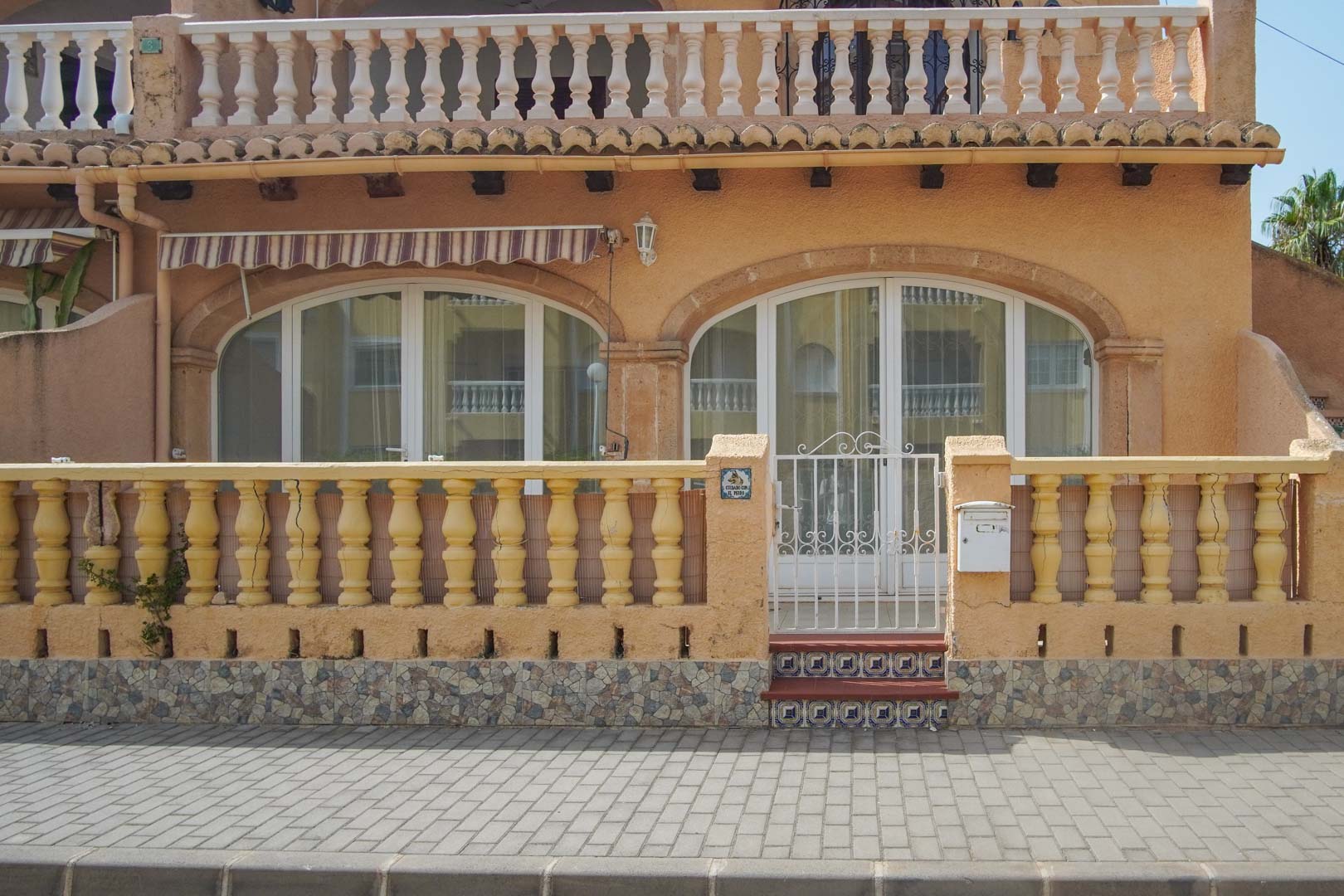 Exterieur van de begane grond met klassieke balustrade, wit metalen toegangshek en boogvormige glazen deuren die toegang bieden tot het terras.