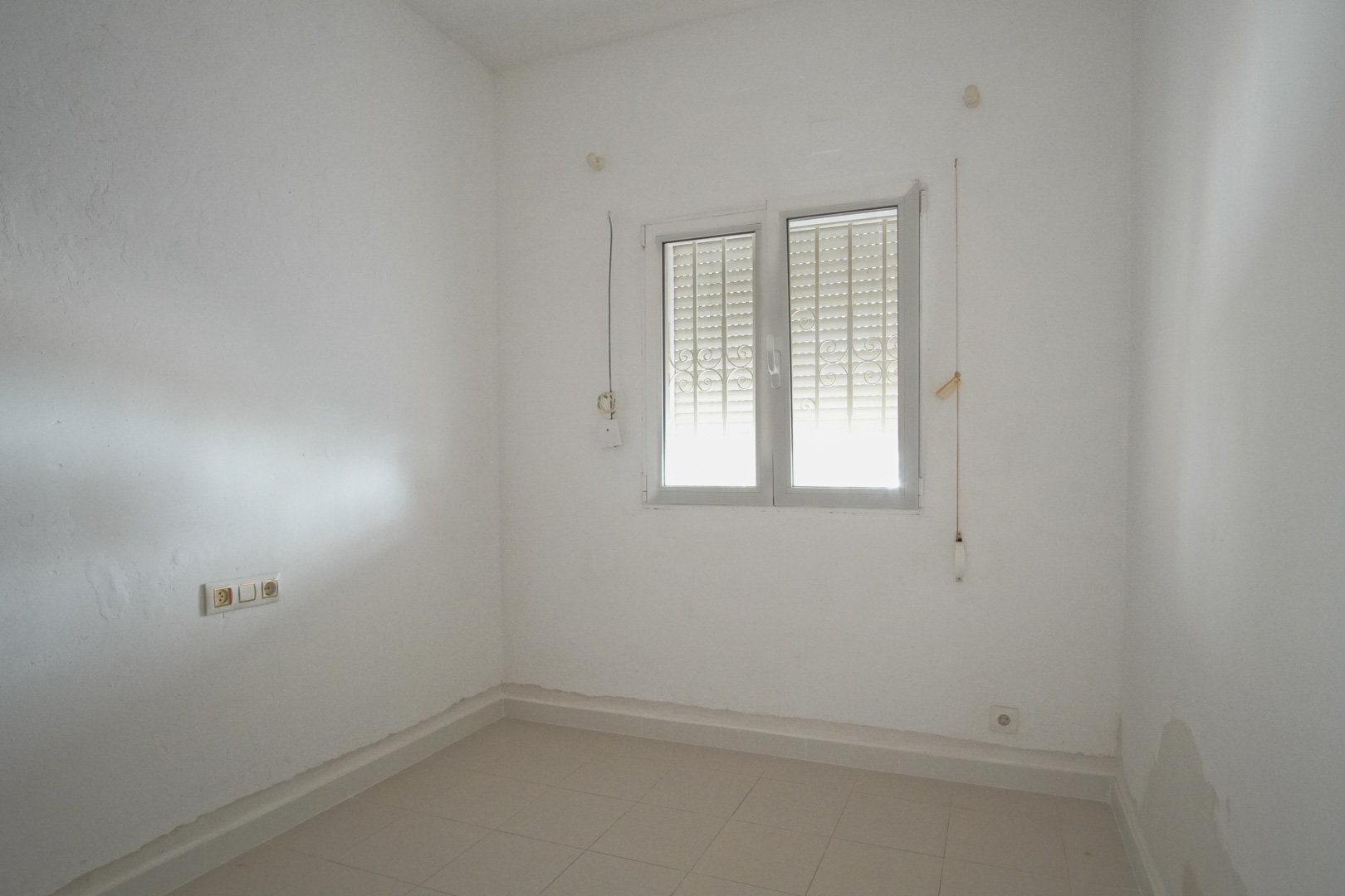 Lichte kamer met witte muren, tegelvloer en een dubbel raam voorzien van veiligheidstralies en geïntegreerde rolluiken.
