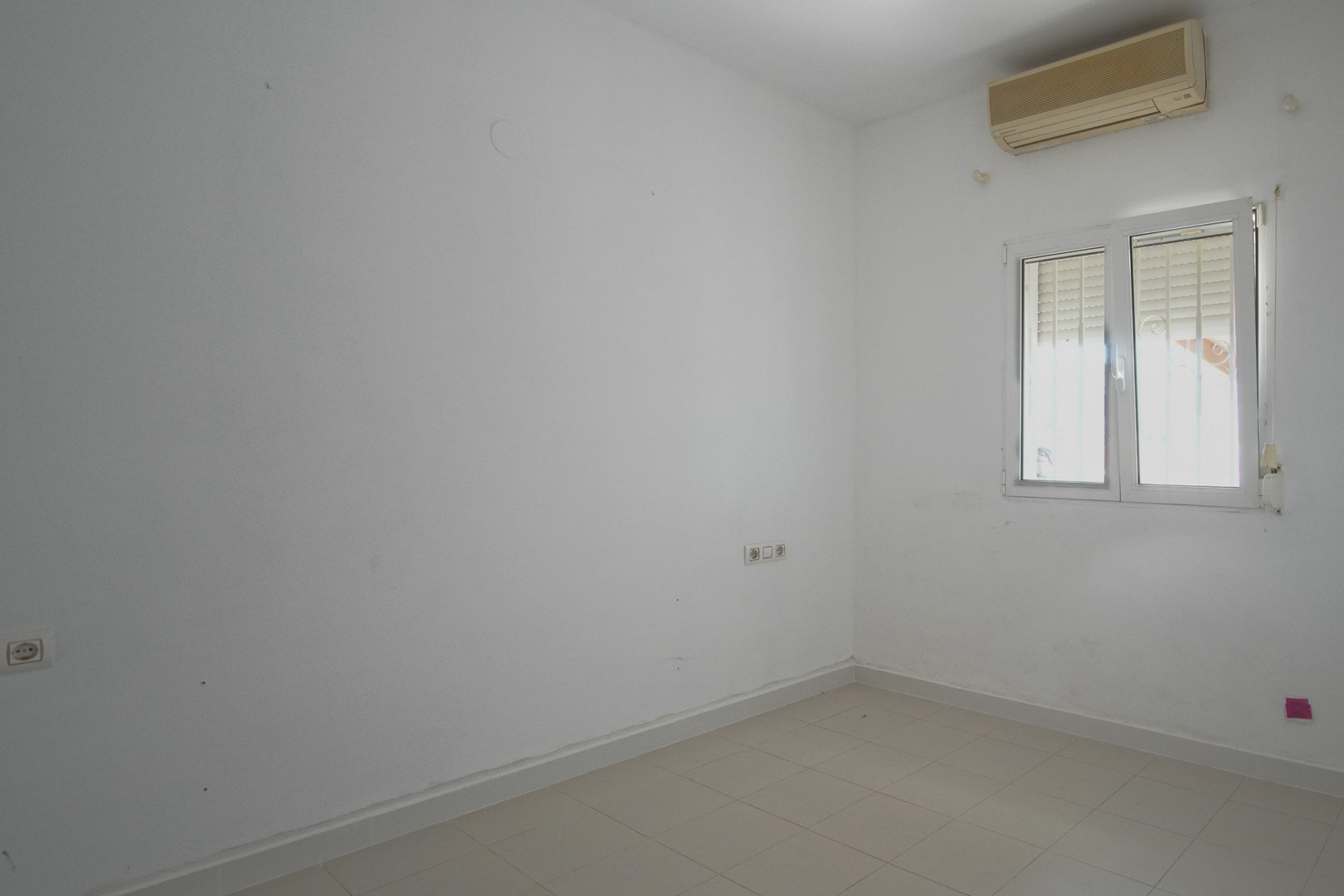 Lichte slaapkamer met witte muren, tegelvloer en een wandgemonteerde airconditioning. Voorzien van een PVC-raam met rolluik en diverse stopcontacten.