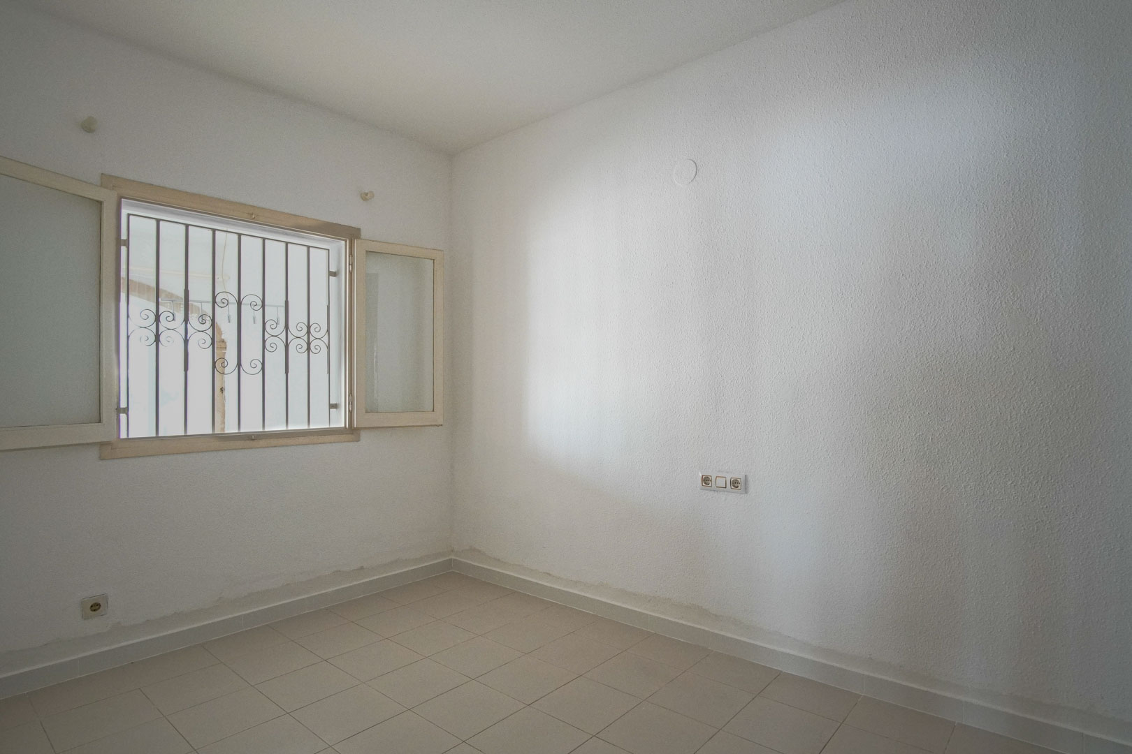 Lichte slaapkamer met witte structuurwanden, lichtgekleurde tegelvloer en een buitenraam voorzien van decoratieve veiligheidstralies.