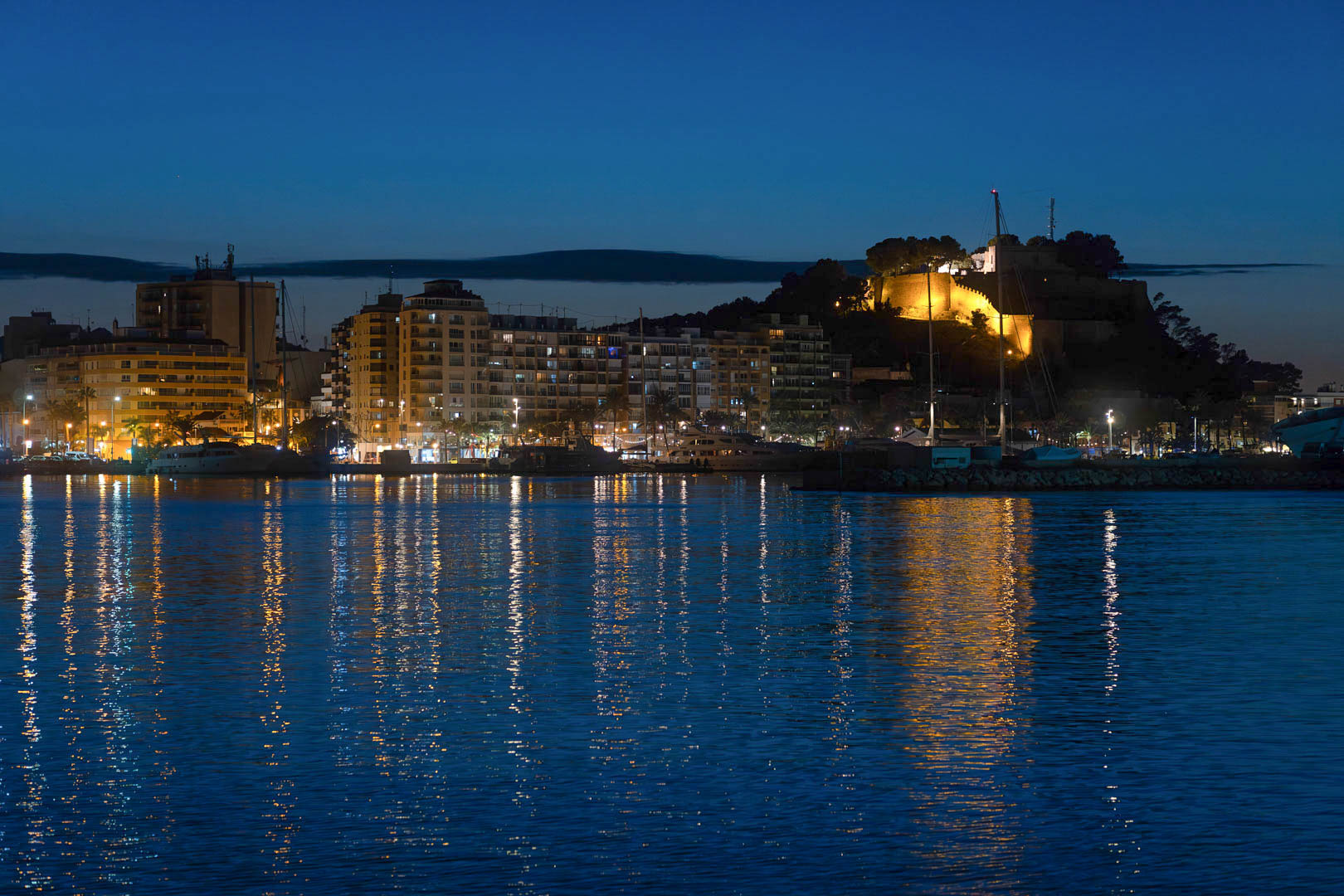 Nachtzicht op de haven van Dénia met het verlichte historische kasteel en de skyline van de stad weerspiegeld in het rustige zeewater.