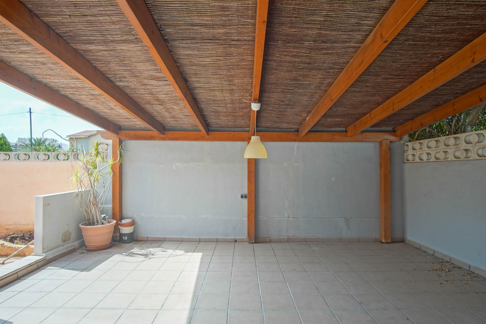 Overdekt buitenterras met een houten balkenstructuur, rieten dakbedekking, tegelvloer en muren met decoratieve windblokken.