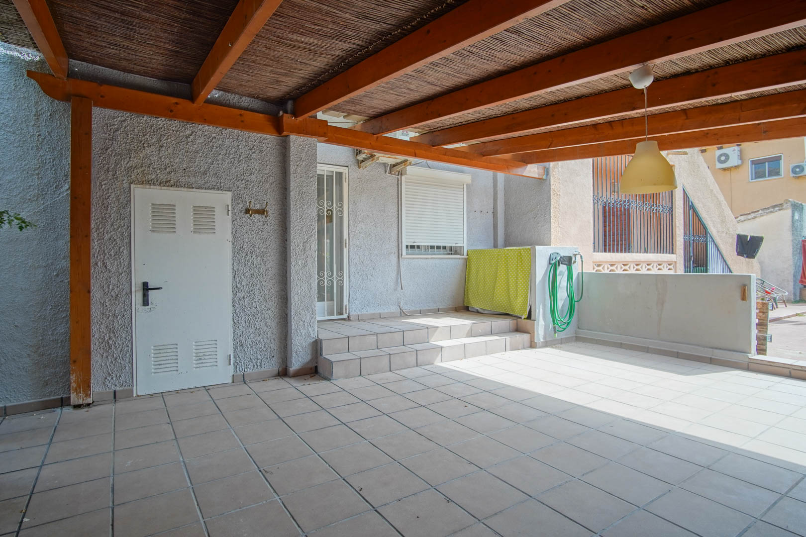Overdekt buitenterras met een houten pergola en rietbedekking, betegelde vloer en directe toegang tot de woning via treden en een witte beveiligingsdeur.