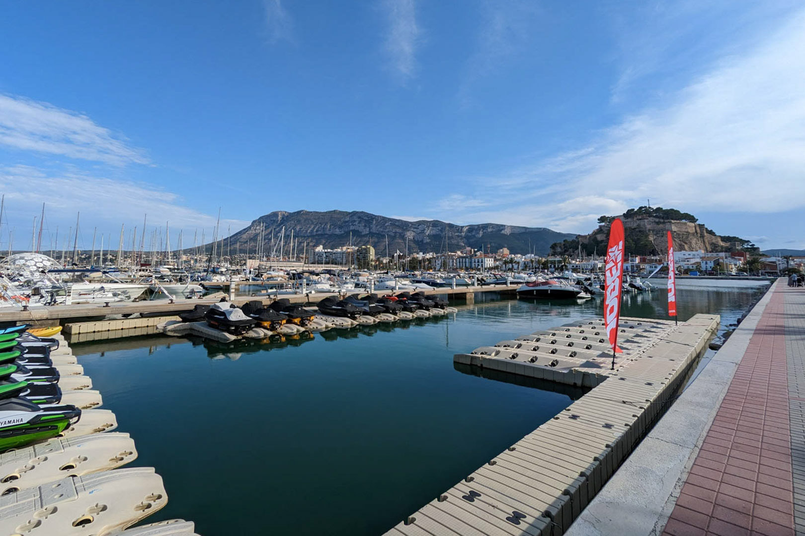 Panoramisch uitzicht op de jachthaven van Dénia, met het historische kasteel en het Montgó-massief op de achtergrond onder een heldere hemel.