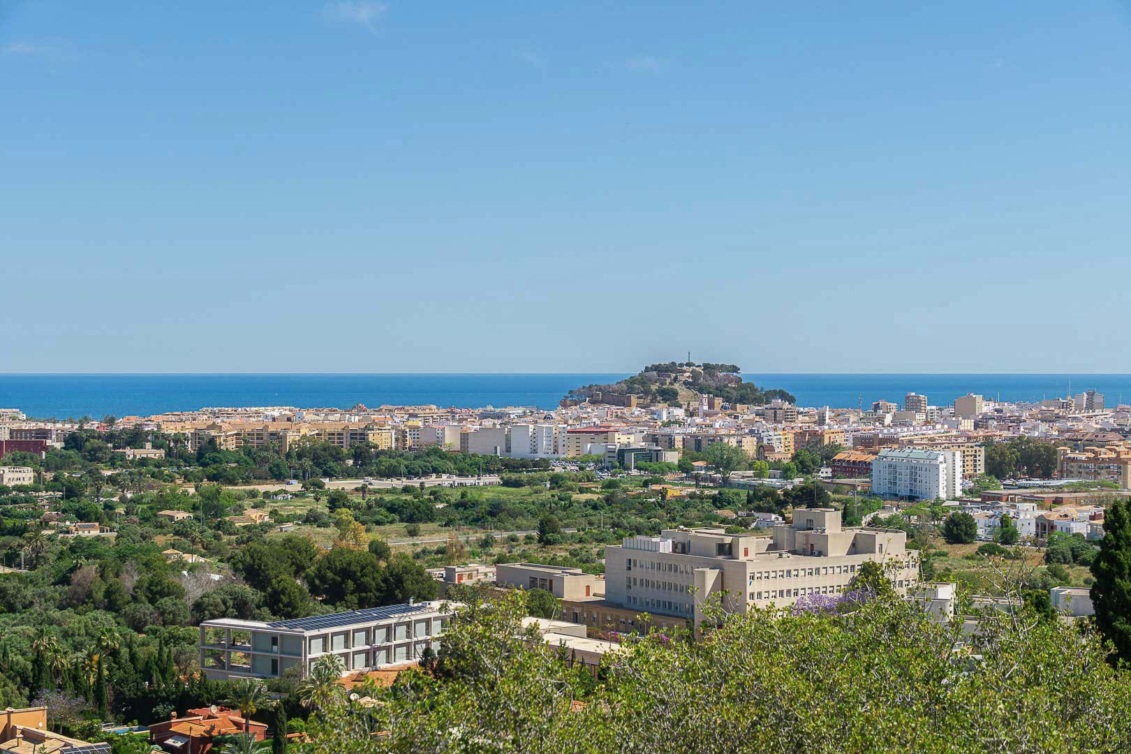 Panoramisch uitzicht op Dénia met het historische kasteel op de heuvel, de Middellandse Zee aan de horizon en het stedelijke landschap omgeven door groen.