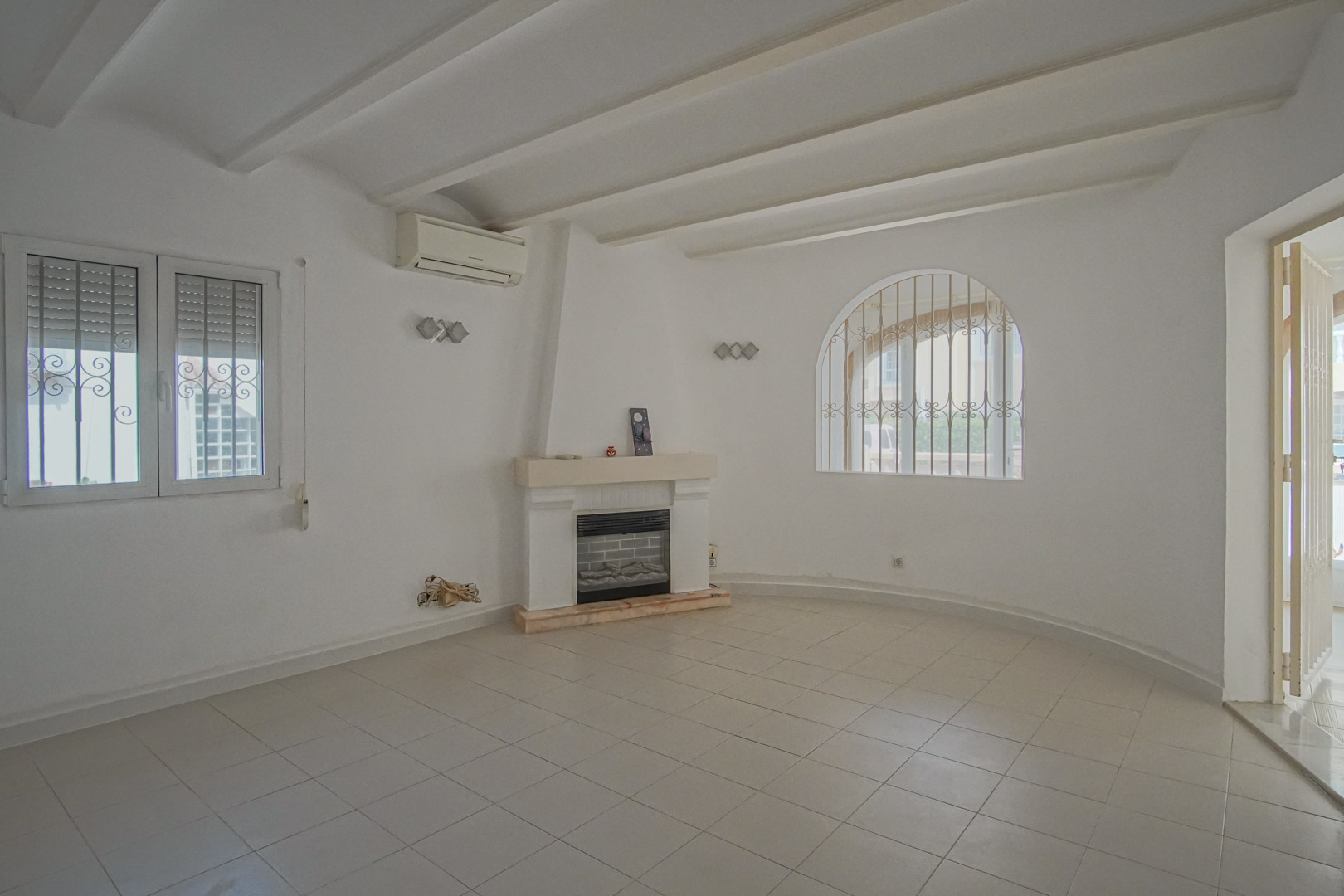 Woonkamer met centrale open haard, wit balkenplafond en een boogvenster met sierhekwerk. Voorzien van airconditioning en een lichte tegelvloer.