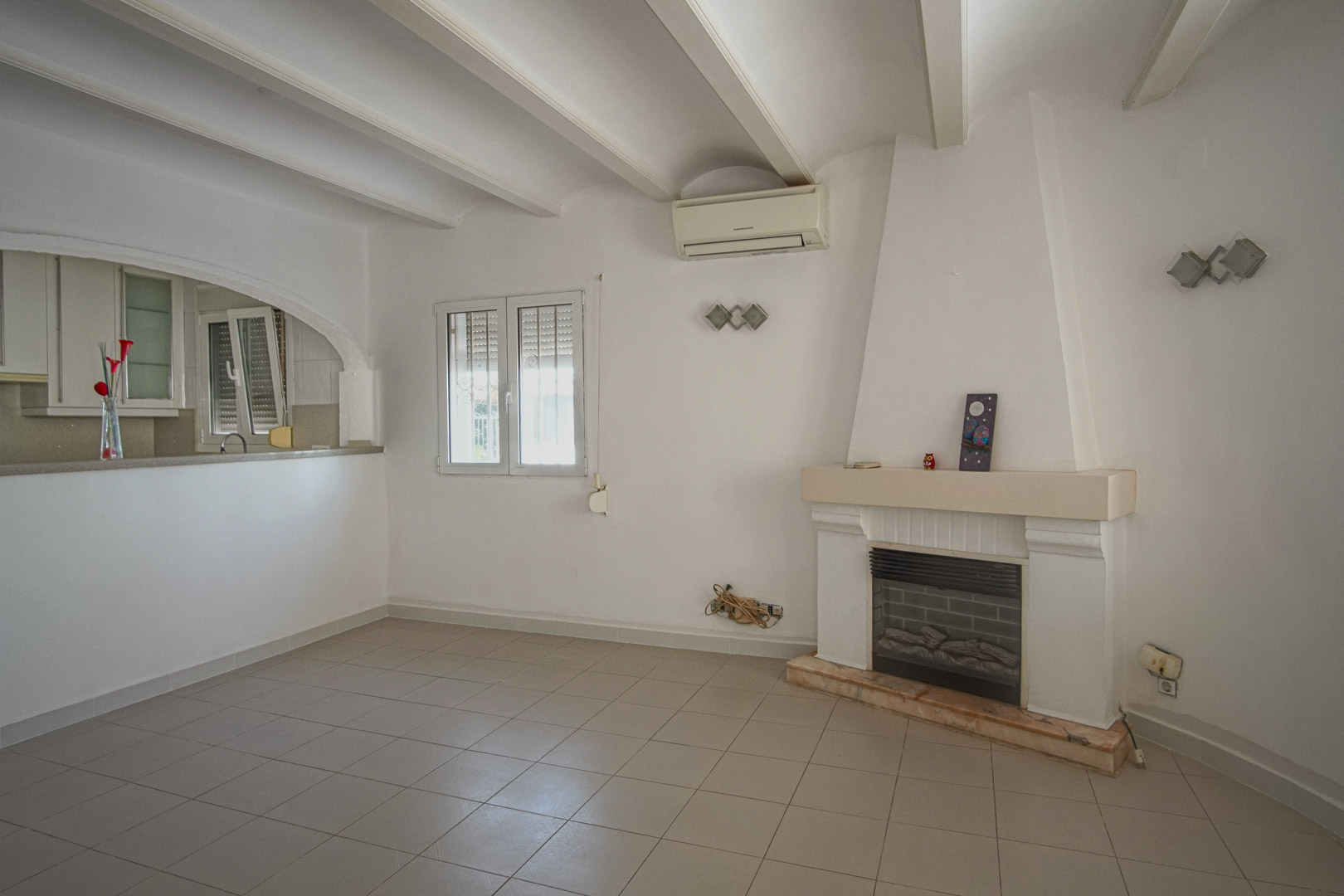 Woonkamer met open haard, wit balkenplafond en airconditioning. Een gewelfde opening verbindt de ruimte met de halfopen keuken.