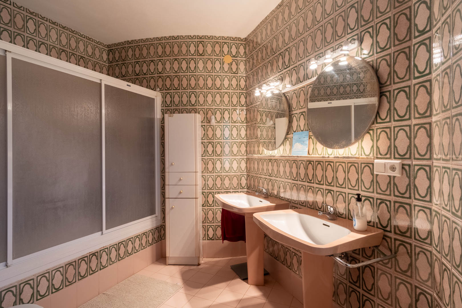 Badkamer in retrostijl met dubbele wastafels, verlichte ronde spiegels en groen gedecoreerde wandtegels. Inclusief inloopdouche met matte schuifdeuren en een bergkast.