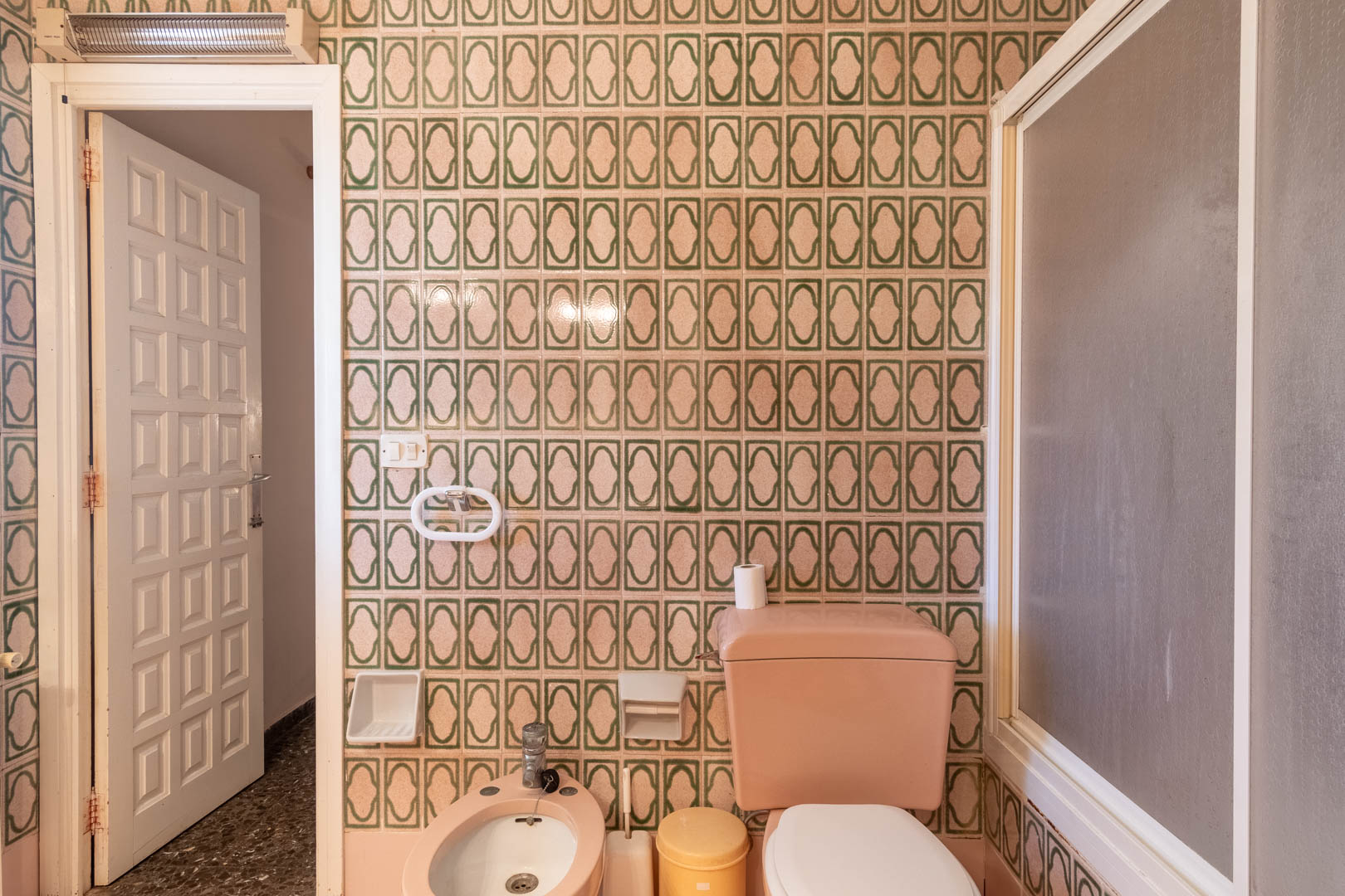 Badkamer met groen gedessineerde wandtegels, roze porseleinen sanitair inclusief bidet en toilet, en een witte paneeldeur.