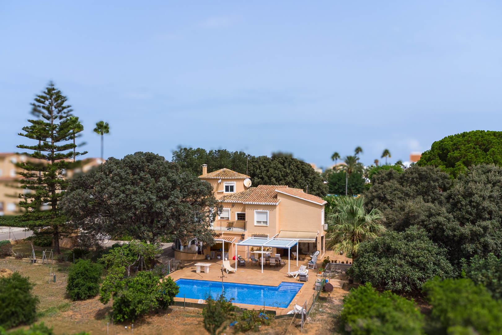 Buitenaanzicht van de villa met rechthoekig zwembad, terras met witte pergola en eethoek, omgeven door volgroeide mediterrane bomen en beplanting op een vlak perceel.