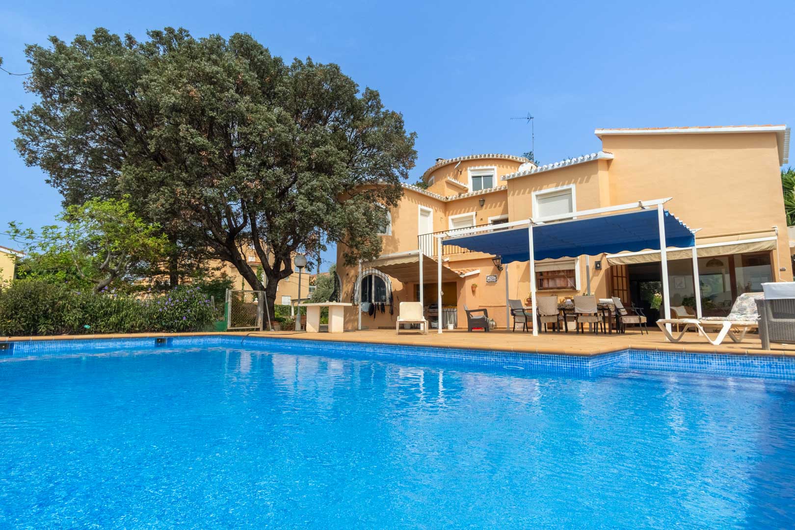 Buitenaanzicht van het privézwembad met ruim zonneterras, de mediterrane villa en een schaduwrijke eethoek onder een blauw zonnescherm.