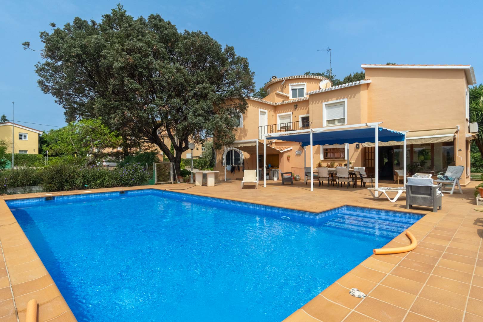 Buitenaanzicht van het rechthoekige zwembad met een terras van terracotta tegels, de mediterrane gevel en een schaduwrijk eetgedeelte onder een blauw zonnescherm.