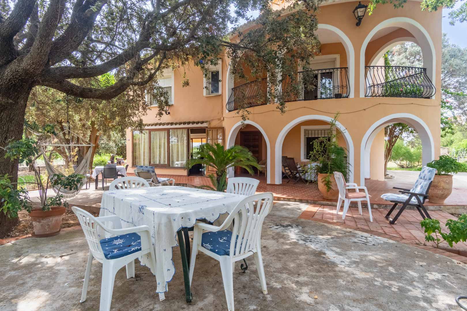 Mediterrane gevel met witte boogvormige veranda's, ruime terrassen in de schaduw van volwassen bomen en buitenruimtes om te eten op terracotta tegels.