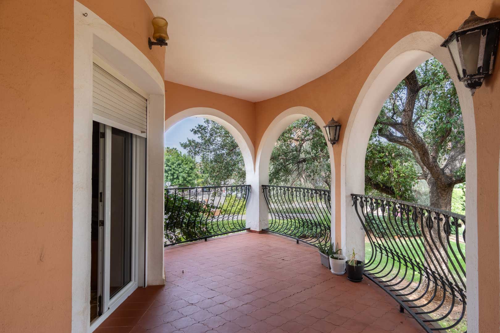 Overdekt terras met mediterrane bogen, gebogen smeedijzeren balustrades en een terracotta tegelvloer met uitzicht op de volgroeide tuin.