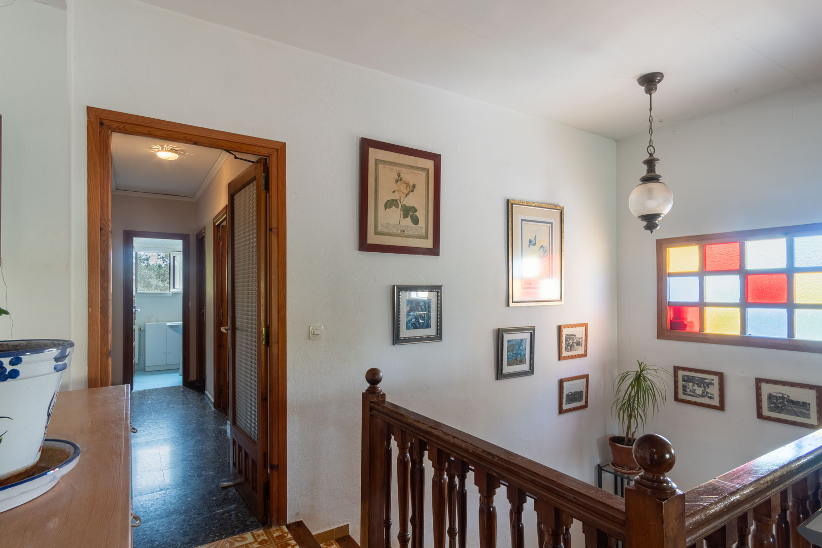 Overloop op de bovenverdieping met gedraaide houten balustrade, kleurrijk glas-in-loodraam en gang naar de slaapkamers met natuurhouten deurkozijnen.