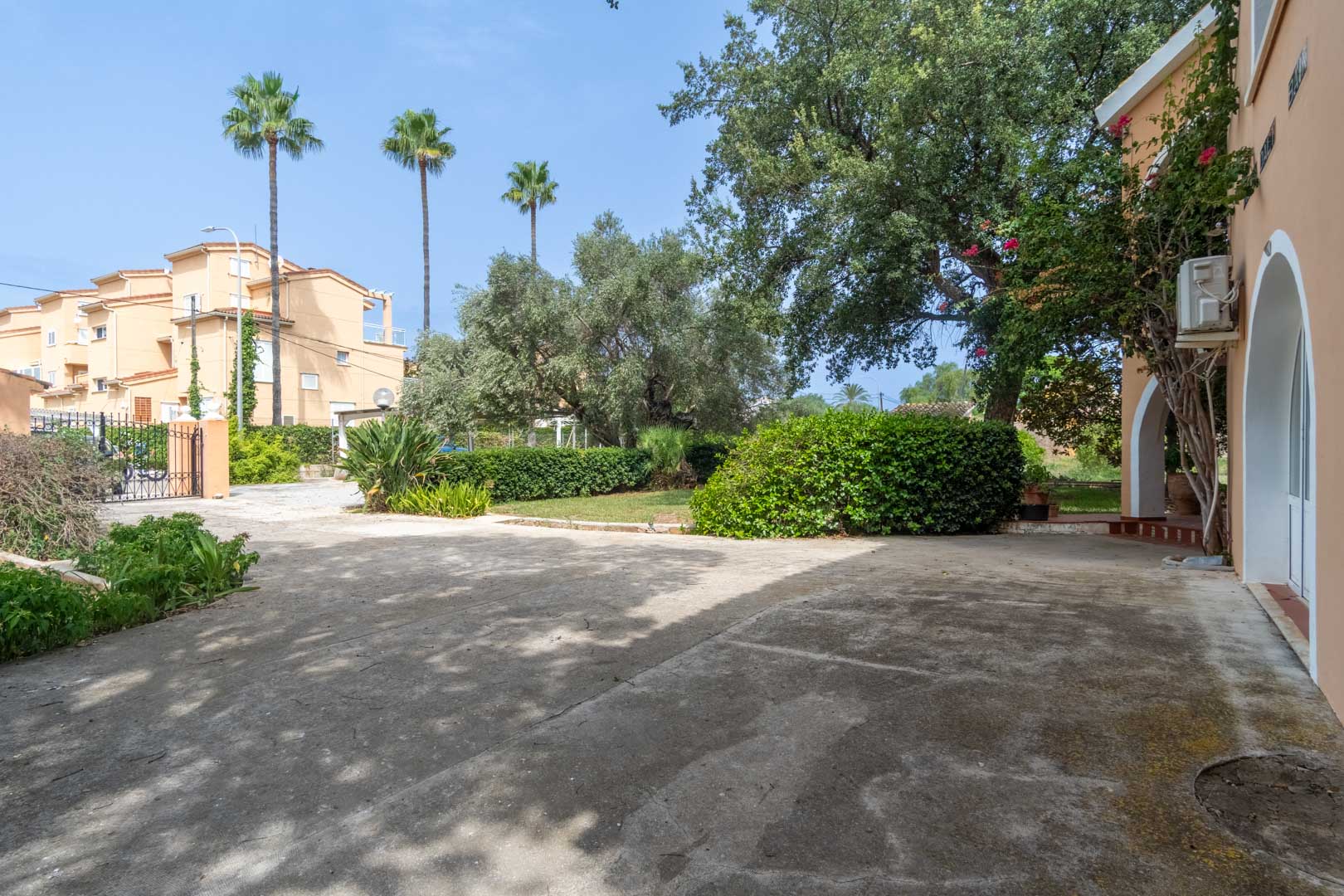 Ruime geplaveide oprit en entree met smeedijzeren poort, omgeven door mediterrane beplanting, palmbomen en traditionele boogvormige architectuur.