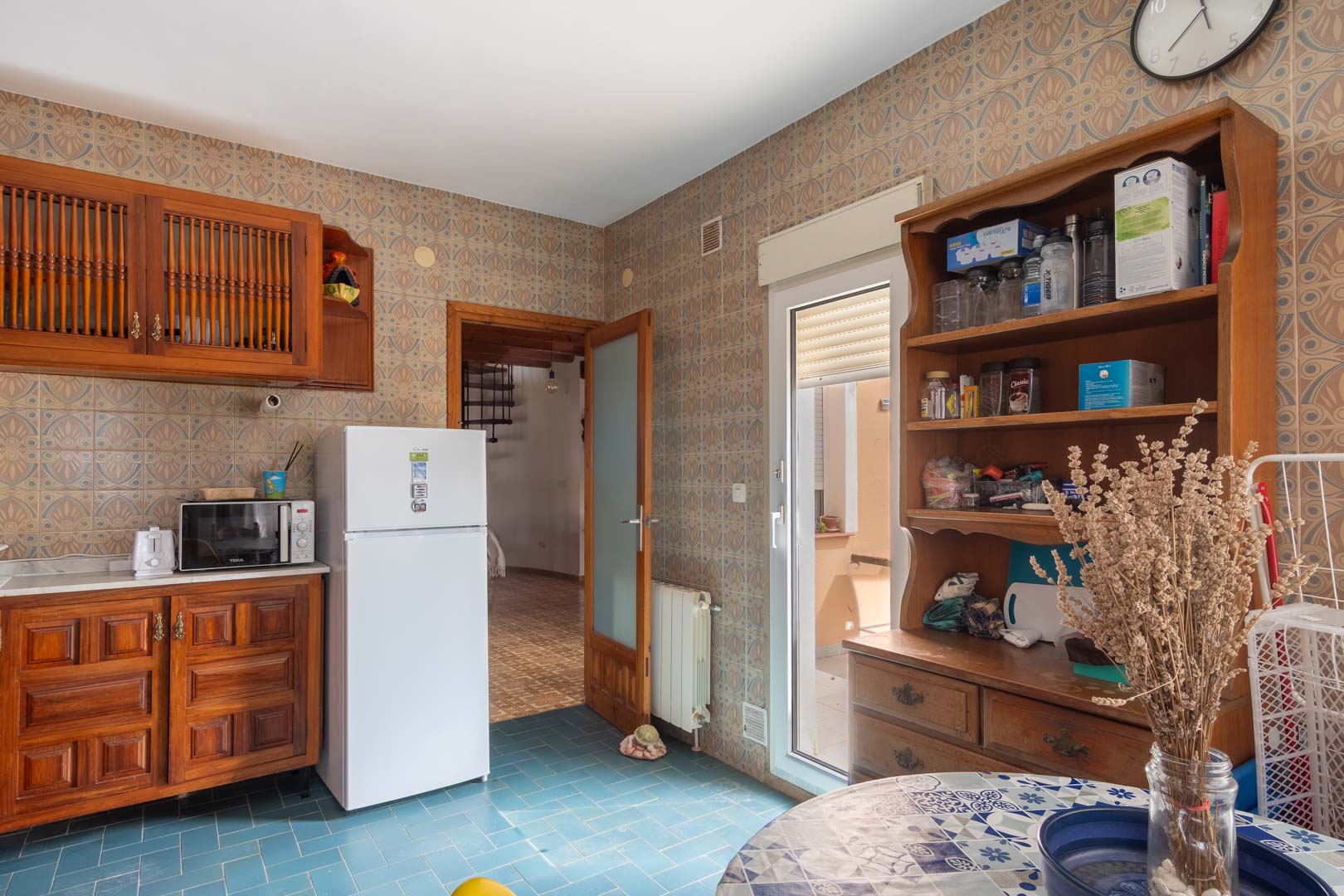 Rustieke keuken met houten kasten, blauwe tegelvloer en gedecoreerde wanden. Voorzien van een glazen deur naar buiten en doorgang naar de naastgelegen woonkamer.