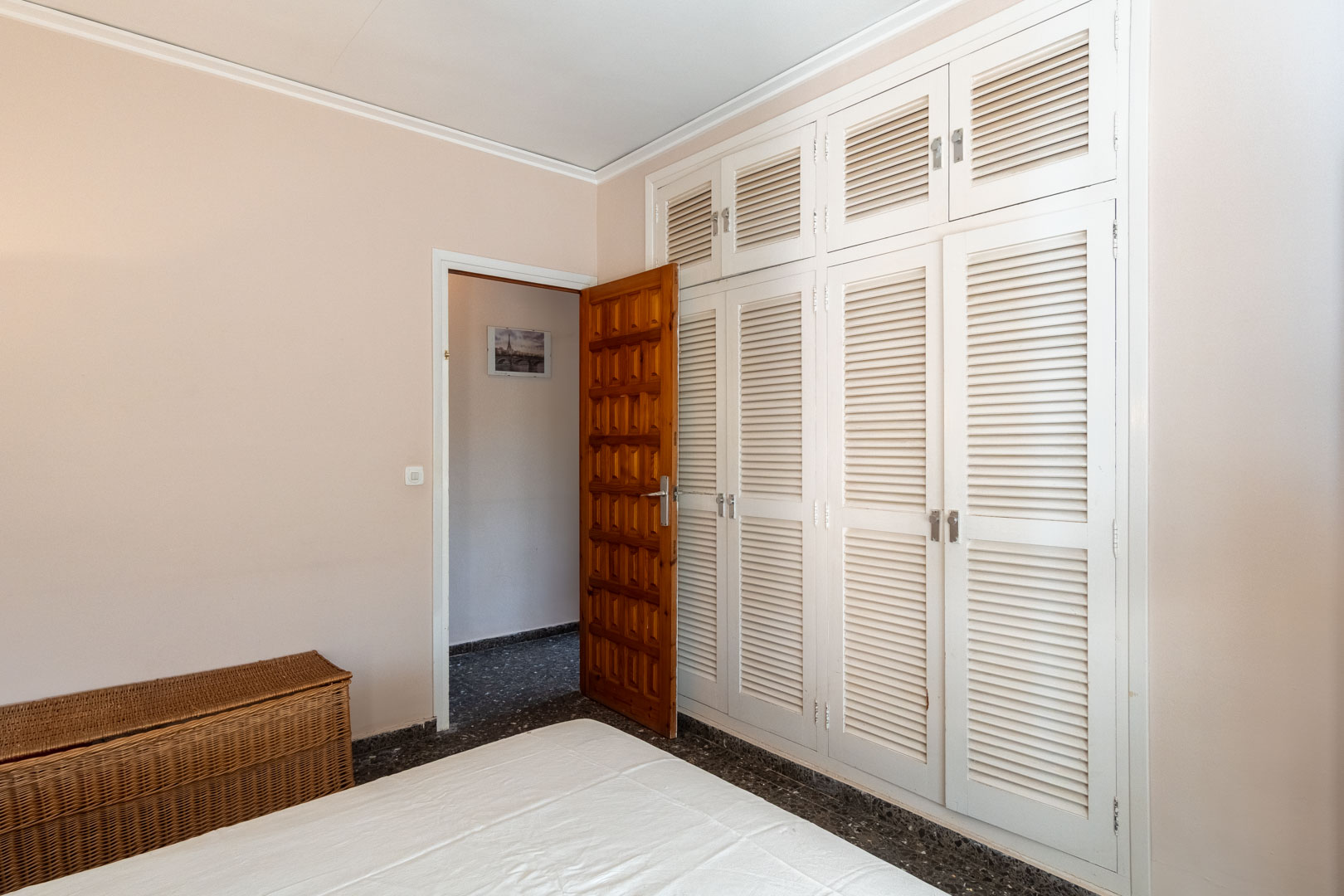 Slaapkamer met een grote inbouwkast met witte lamellendeuren, een donkere terrazzovloer en een massief houten paneeldeur naar de gang.