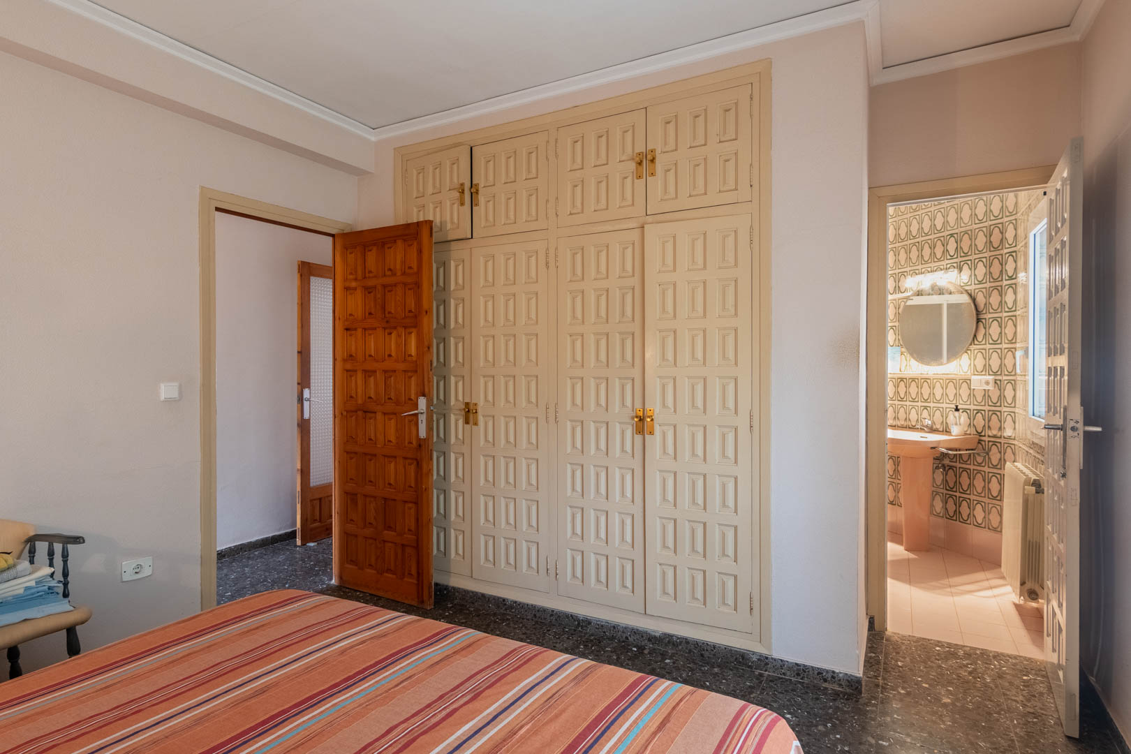 Slaapkamer met een grote ingebouwde kastenwand met paneeldeuren en een en-suite badkamer met geometrische tegels en natuurlijke lichtinval.