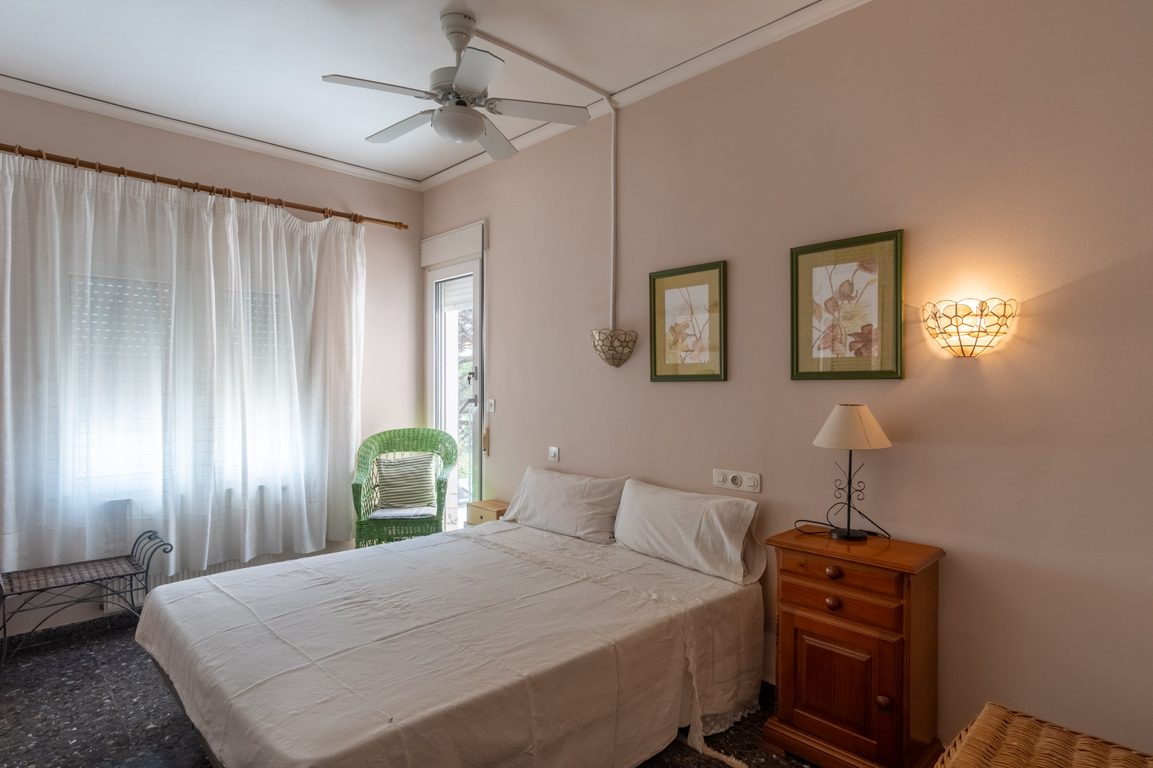 Slaapkamer met tweepersoonsbed, plafondventilator en directe toegang tot buiten. Voorzien van houten meubilair, een terrazzovloer en een decoratieve rotan stoel bij het raam.