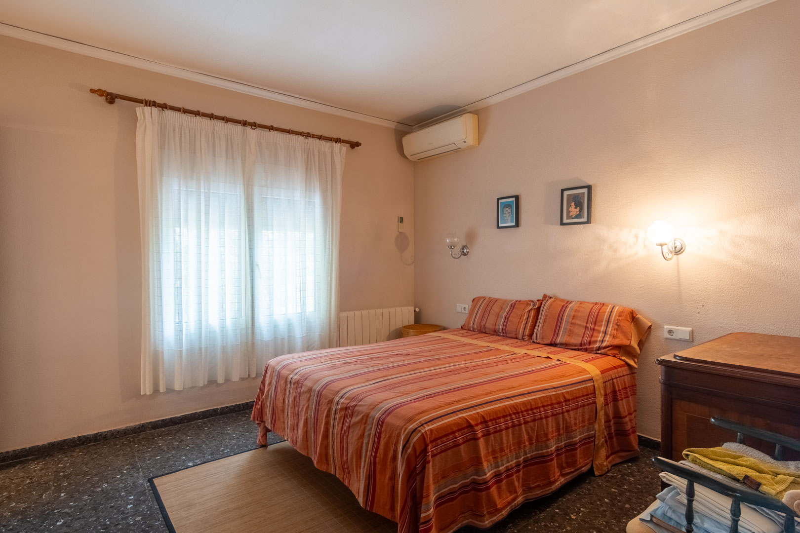 Tweepersoonskamer met donkere terrazzovloer, voorzien van wandairconditioning, een radiator en een raam met witte gordijnen voor natuurlijk lichtinval.