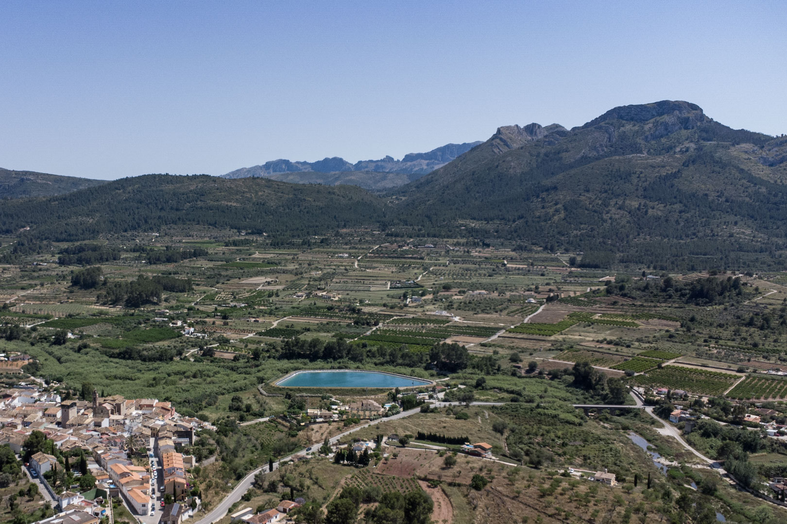 Panoramisch luchtfoto van de Jalón-vallei vanuit Alcalalí, met uitzicht op de landbouwgronden, een turquoise waterreservoir en de omliggende bergen.