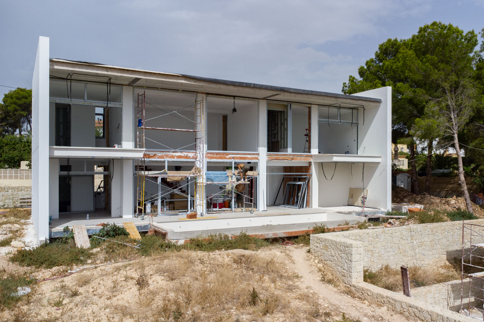 Buitenaanzicht van de villa in aanbouw met de witte gevel van twee verdiepingen, grote raamopeningen en de aanleg van het zwembad.