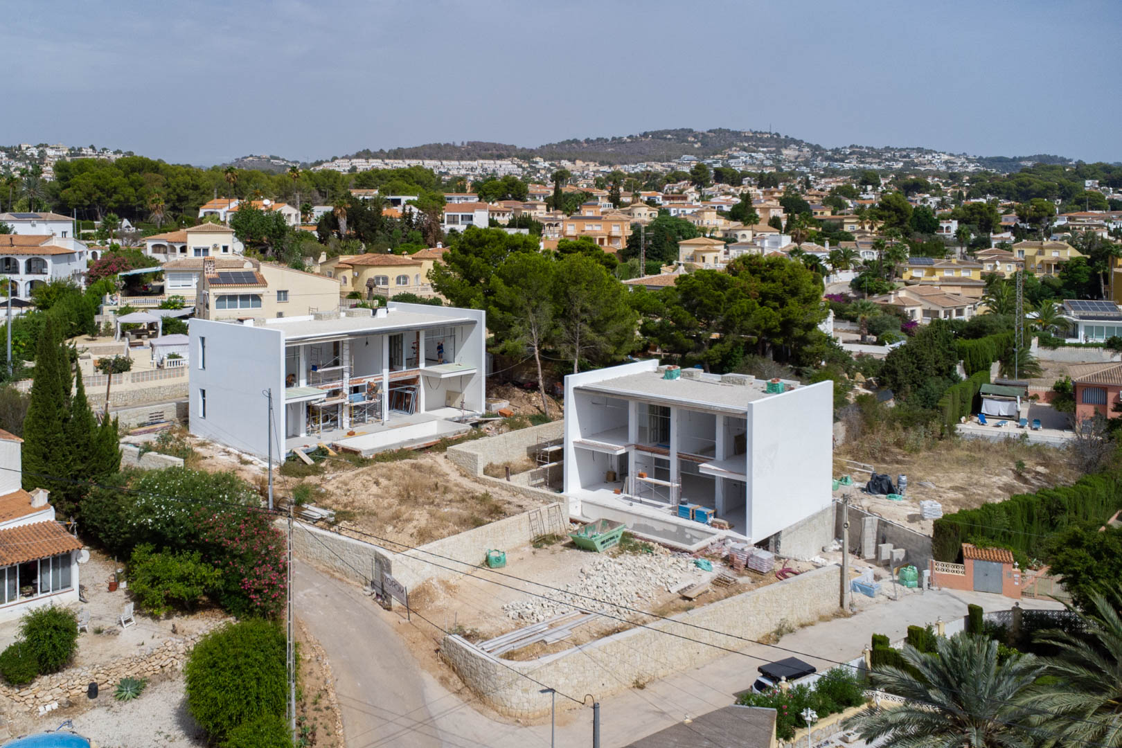 Luchtfoto van de bouwfase van twee moderne villa's in Calpe, met de witte structuren, natuurstenen muren en de nivellering van de percelen.