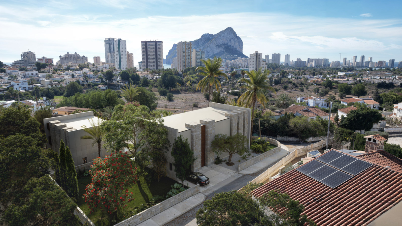 Luchtfoto van de moderne villa met natuurstenen gevels en aangelegde tuin, met uitzicht op de Peñón de Ifach en de skyline van Calpe.